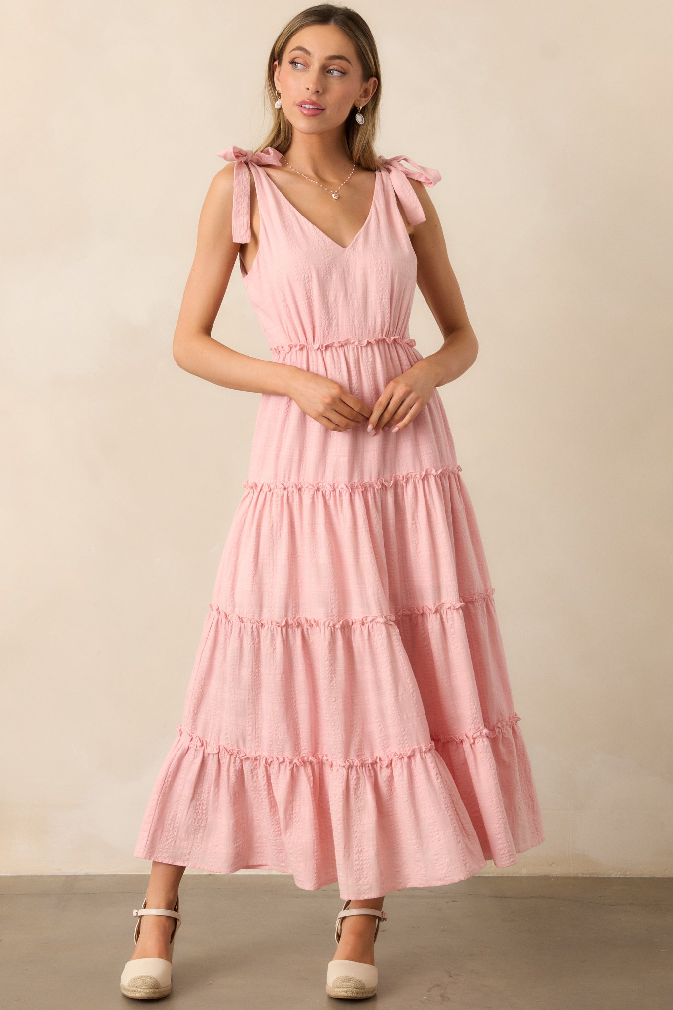 A pink dress with a flowing tiered skirt, v-neckline, and self-tie bow straps, finished with ruffle detailing and made from a lightweight textured fabric.