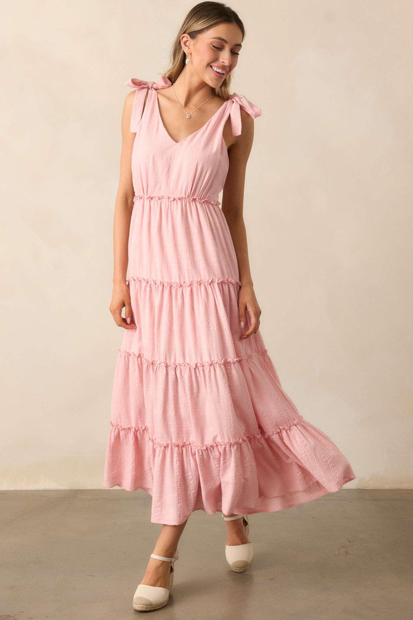 A pink dress featuring a flattering v-neckline, tiered skirt, and self-tie bow straps, with delicate ruffle detailing and a soft textured fabric.