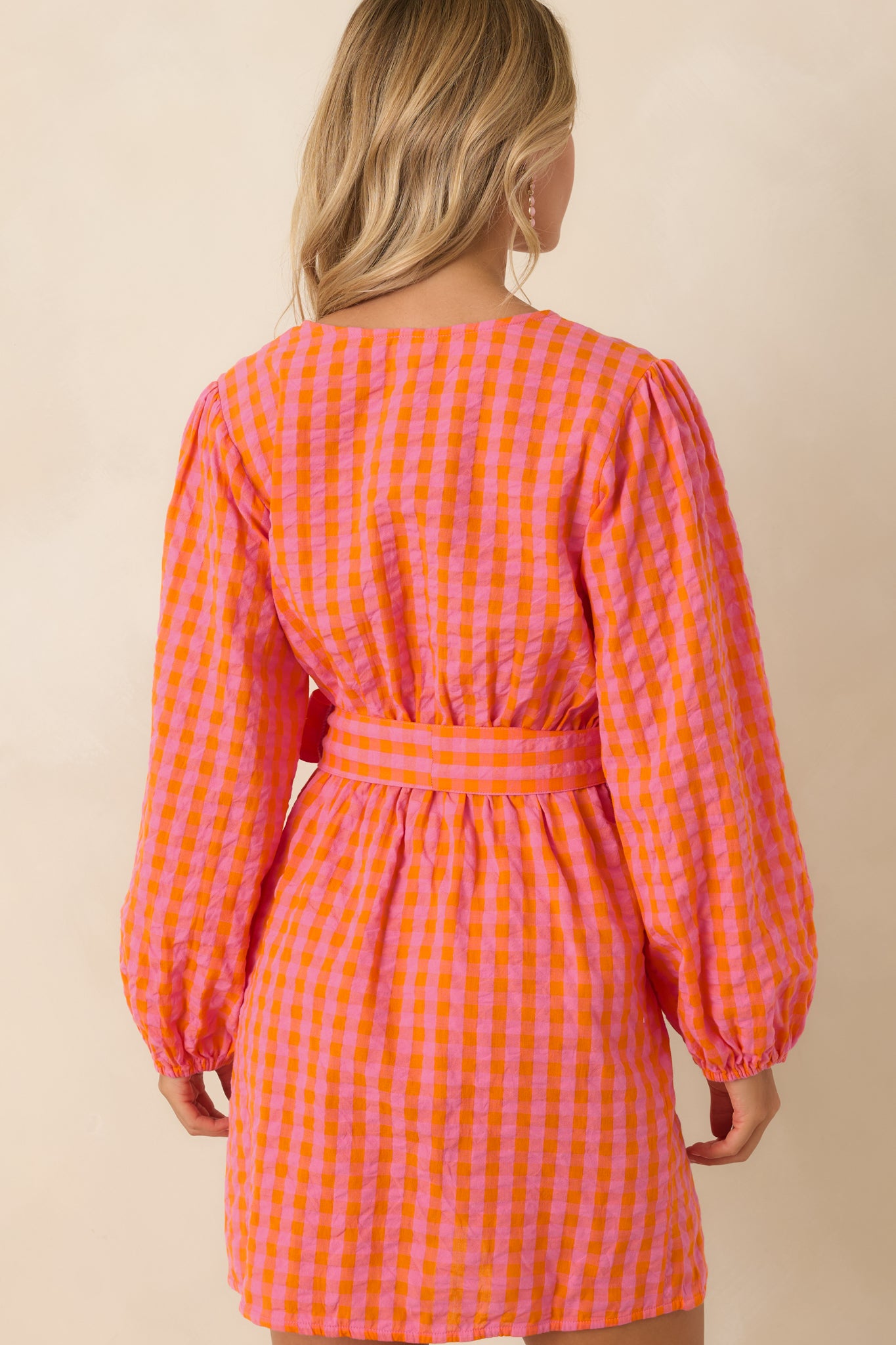 Rear view of the pink and orange cotton gingham wrap mini dress featuring long sleeves, an adjustable tie waist, and a flattering wrap silhouette.