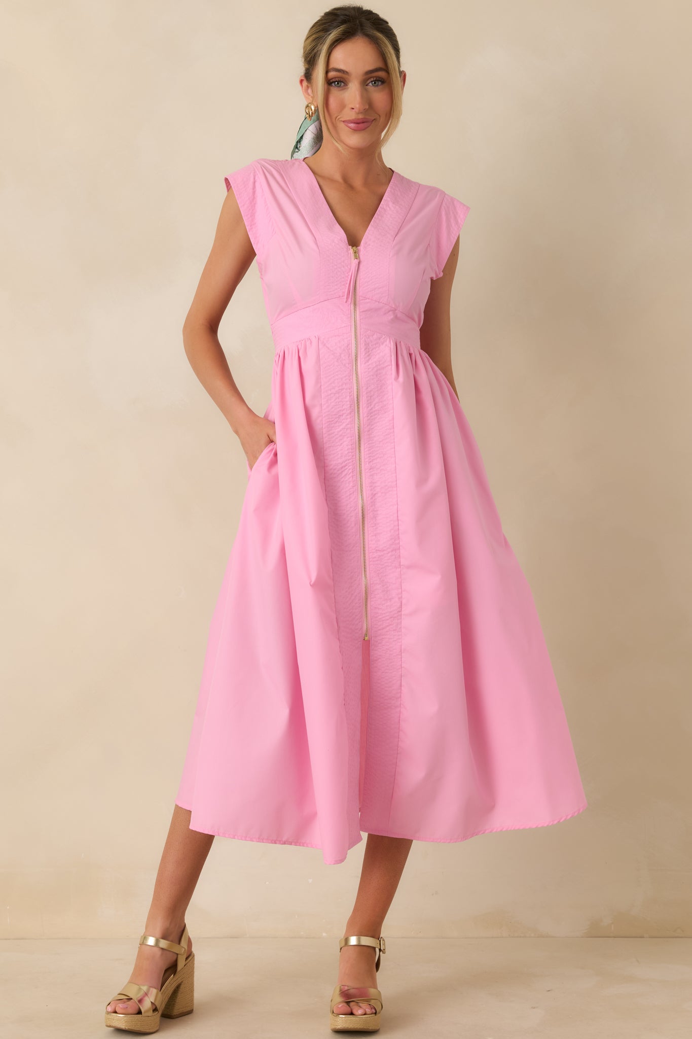 Full body front view of a pink sleeveless midi dress featuring a V-neckline, functional gold-tone front zipper, and a fitted waist flowing into a softly gathered skirt.