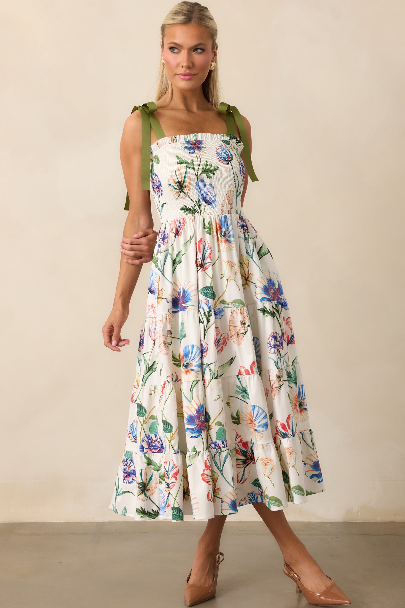 Breezy ivory midi dress with colorful floral print, smocked bodice, and adjustable green bow straps for a whimsical touch.