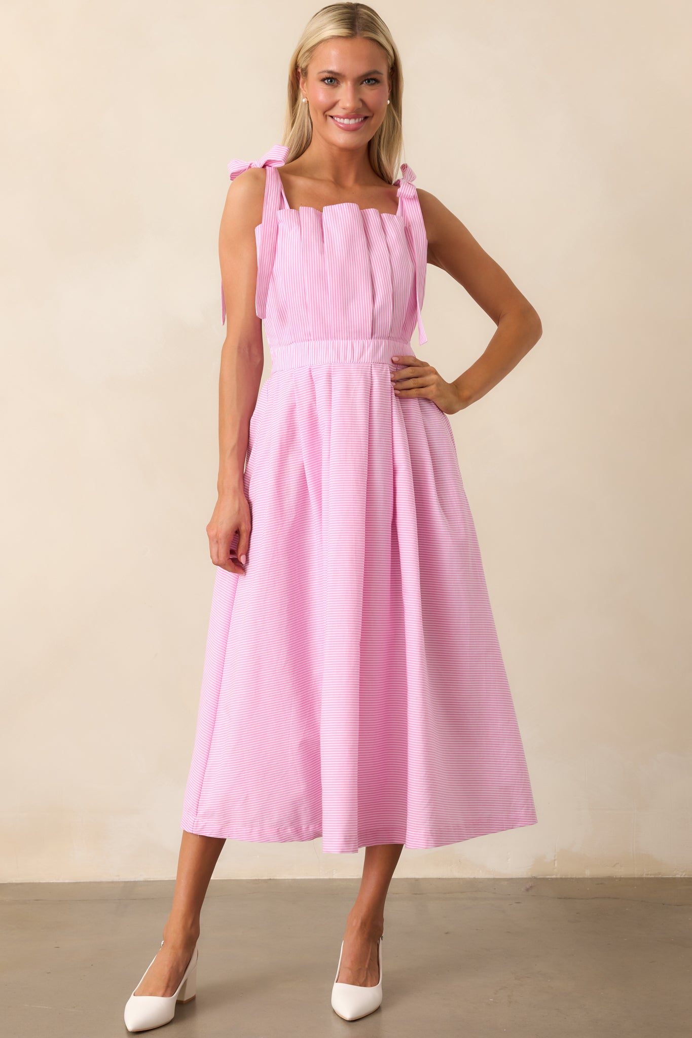 The pink striped midi dress with adjustable self-tie bow straps and a pleated skirt, displayed in a full-length shot.