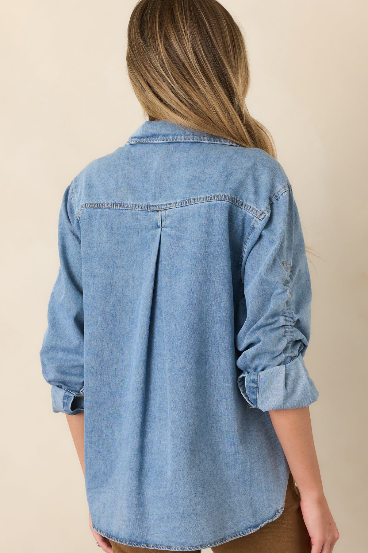 Rear of the light wash denim shirt featuring a back yoke with a center pleat, long sleeves, and a curved hemline.