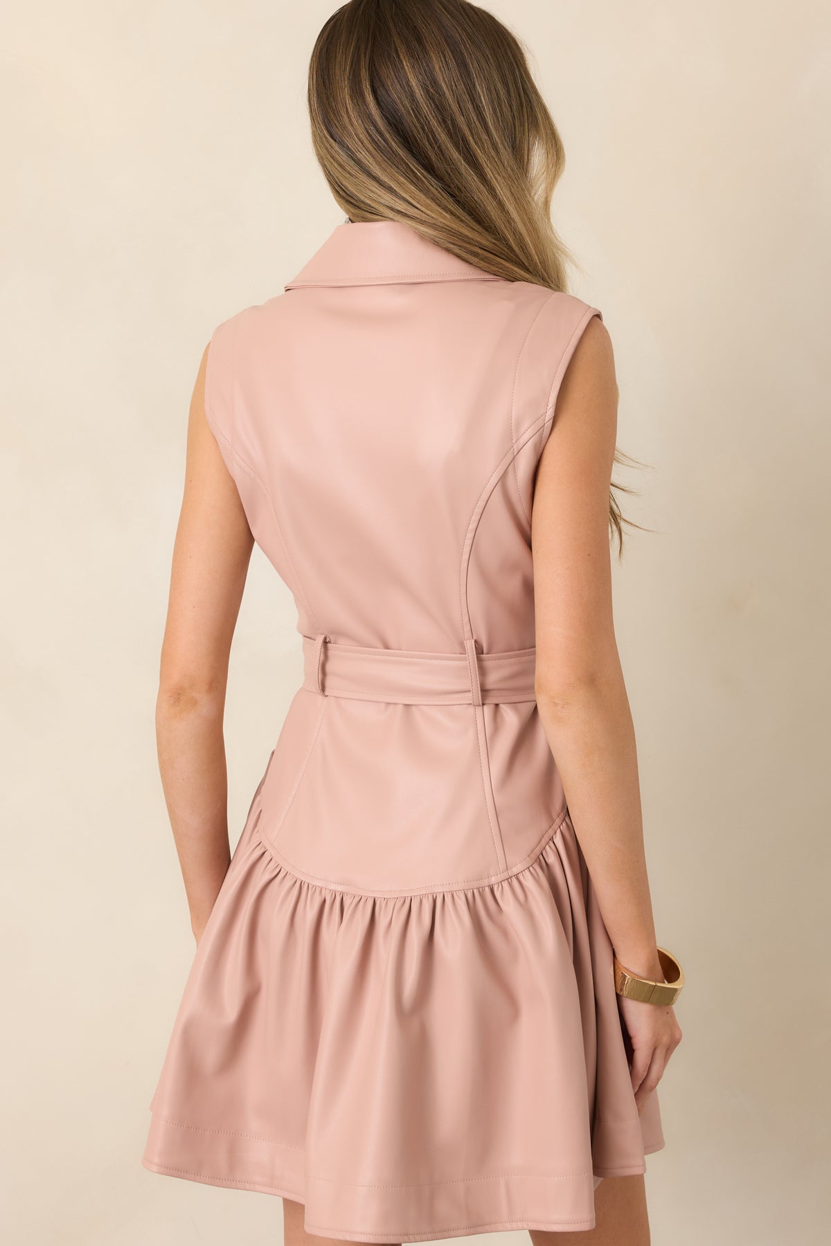 Rear view of the blush pink faux leather mini dress showcasing the sleeveless silhouette, defined waist, matching belt, and gathered skirt.