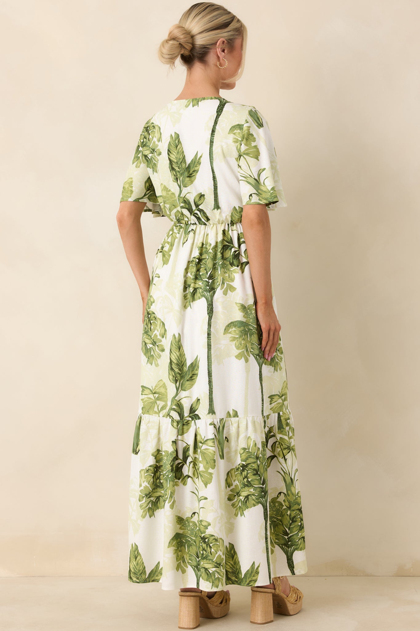 Rear view of ivory and green tropical print maxi dress highlighting flutter sleeves, shaped waistline, and flowing tiered skirt silhouette.