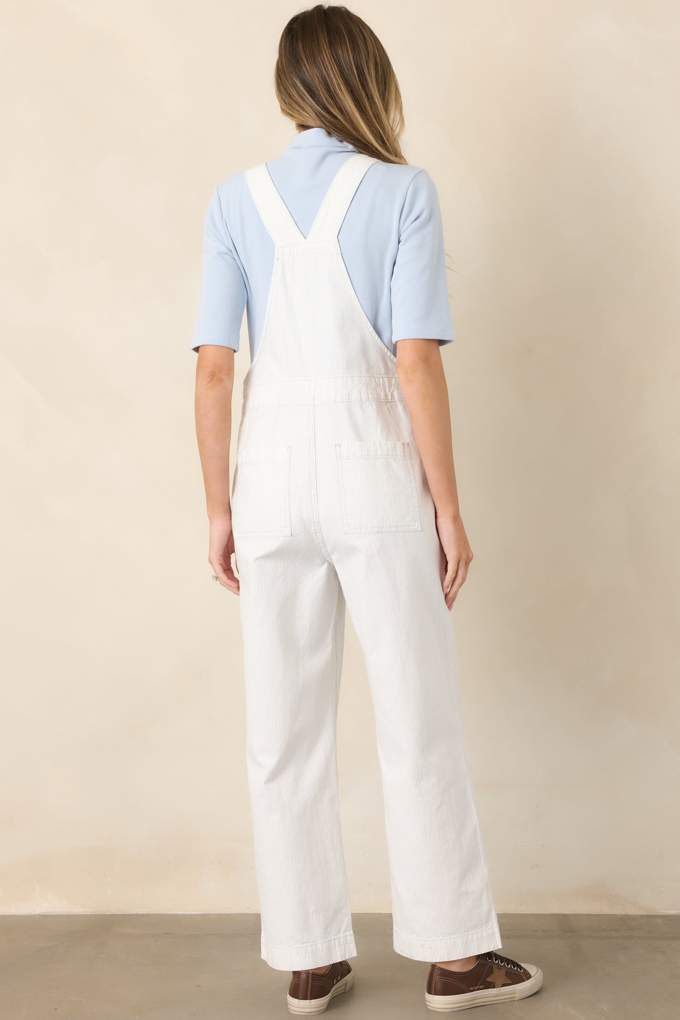 Rear view of the ivory and sky blue striped cotton overalls showcasing the adjustable cross-back straps, side buttons, straight-leg silhouette, and ankle-length hem.
