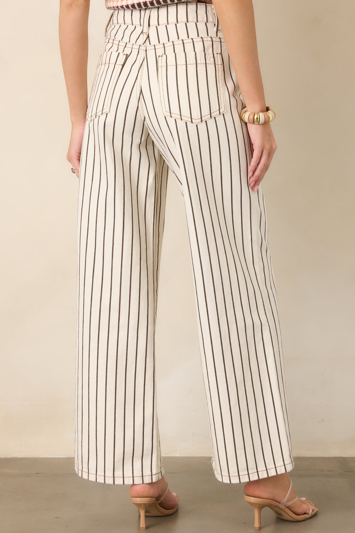 Rear view of the ivory cotton denim pants featuring vertical brown pinstripes, a high-rise waist, back pockets, and relaxed wide legs.
