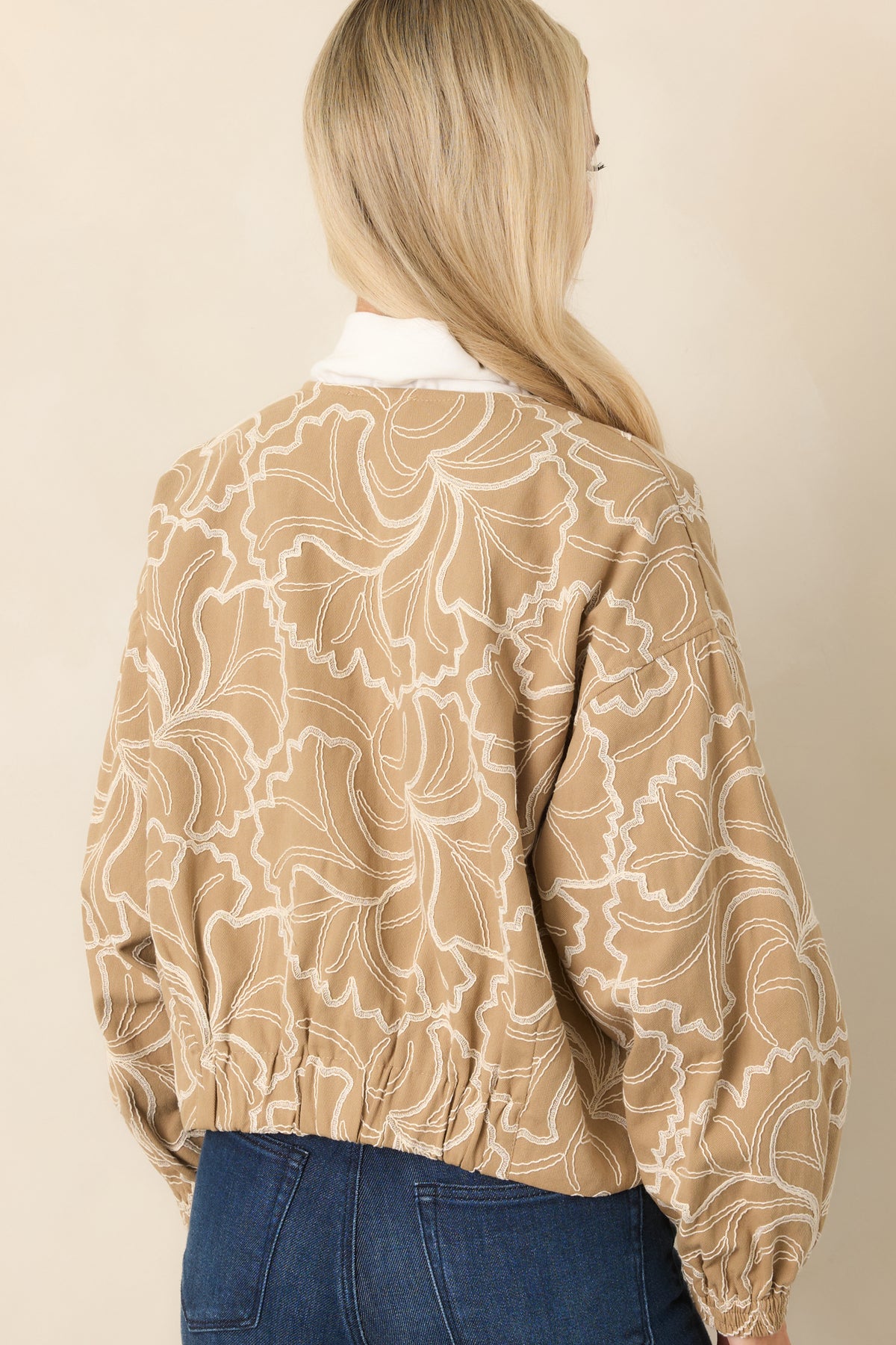 Rear view of the tan cotton jacket featuring an intricate ivory floral embroidered design and relaxed cropped silhouette.