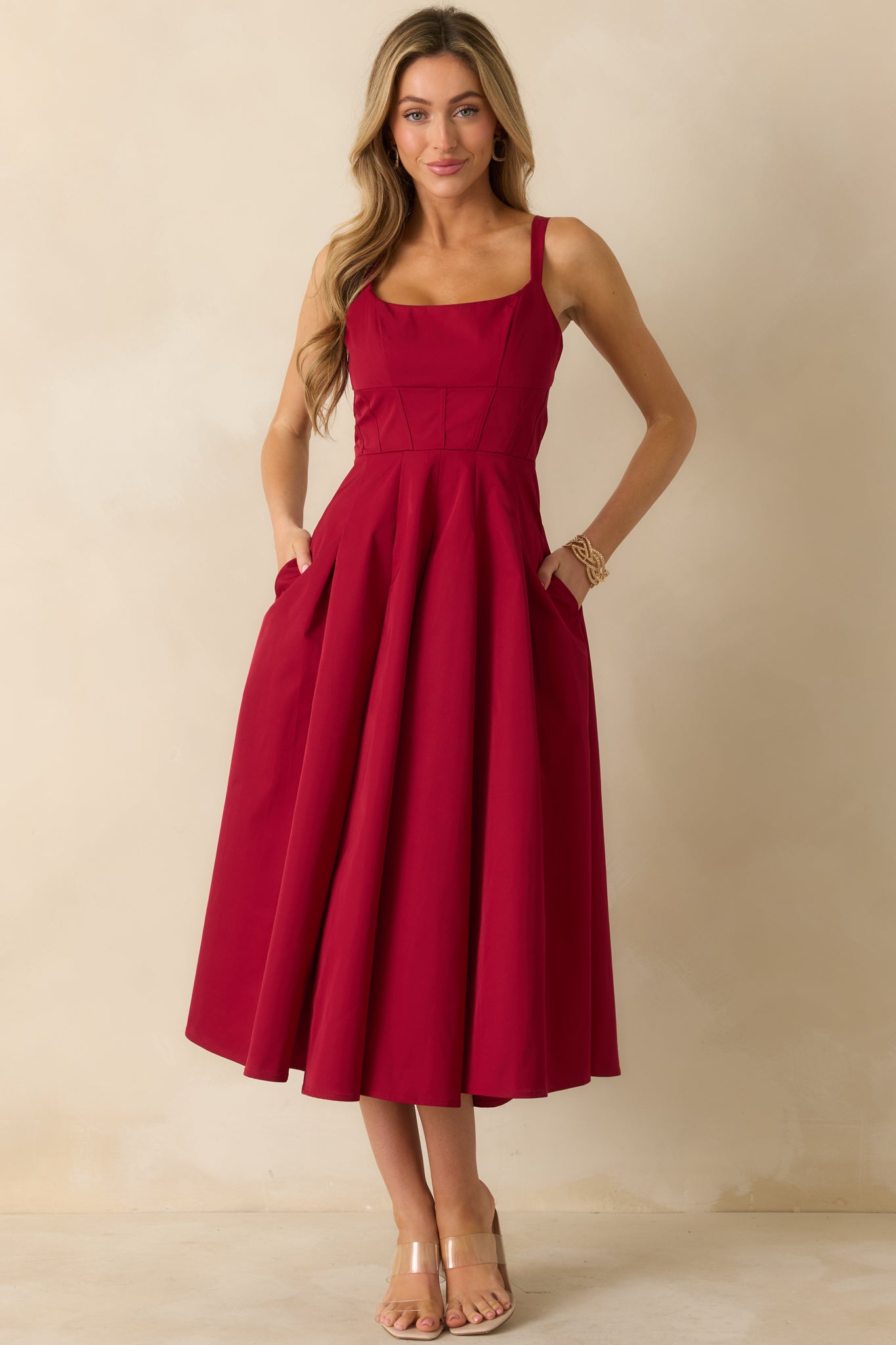 Polished red dress featuring wide straps, seamed bodice detailing, and hidden side pockets within the flowing pleated skirt.