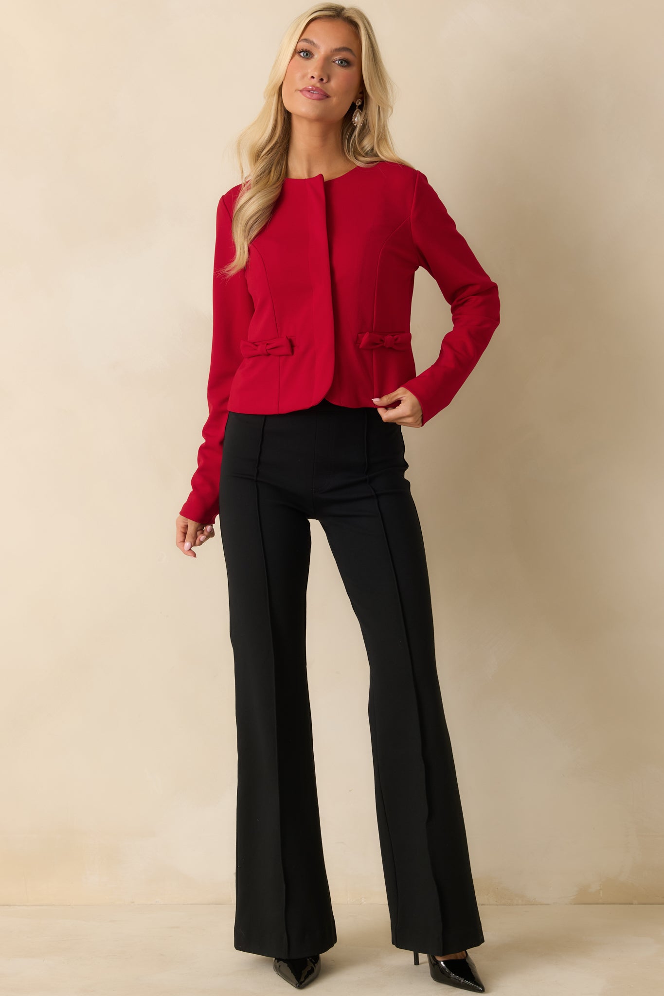 Red jacket with a cropped, tailored silhouette, collarless neckline, long sleeves, and bow-accented faux pockets, shown fully front-facing.