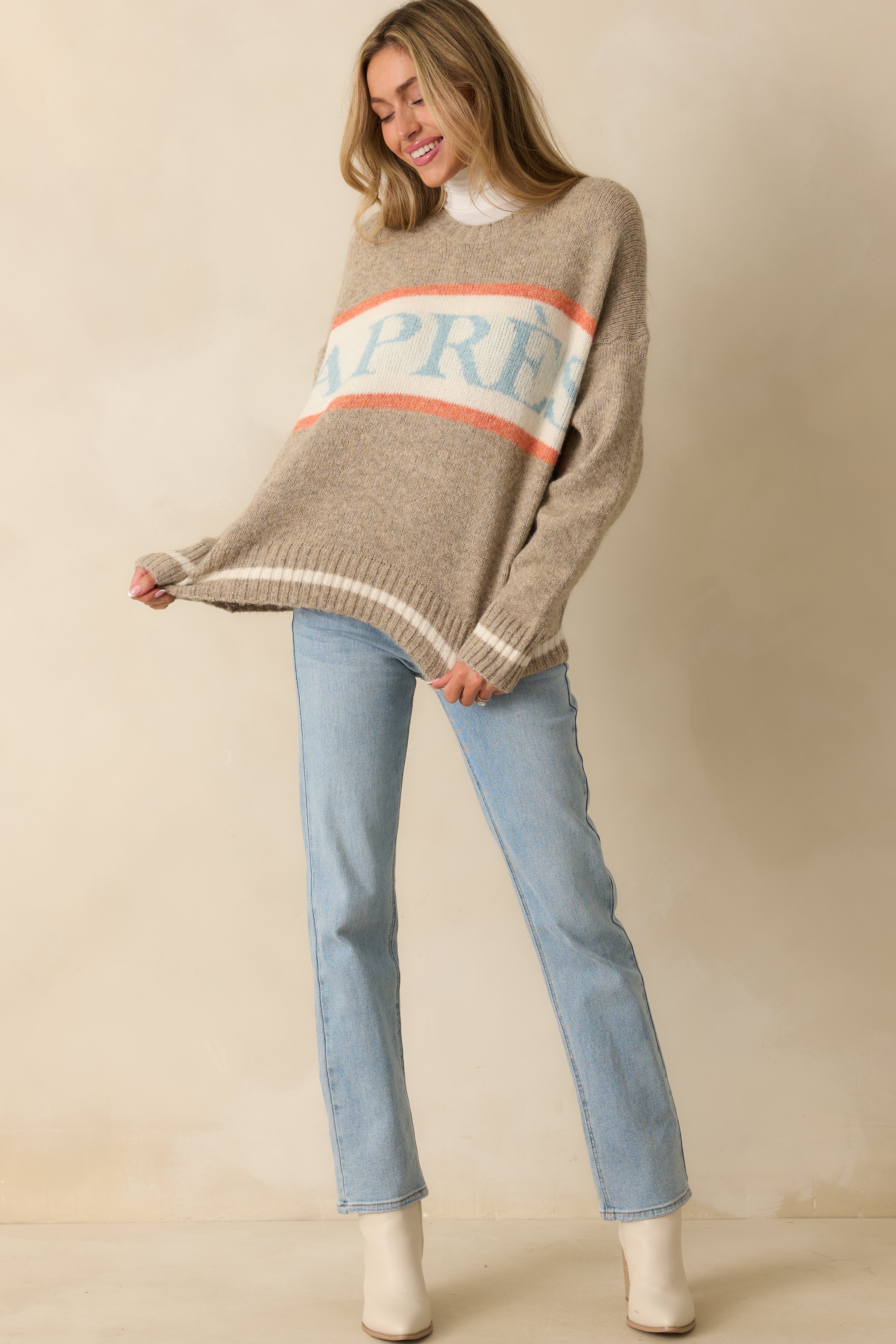 Relaxed taupe knit pullover with long sleeves, ribbed cuffs and hem, and contrasting orange and ivory stripes across the chest.