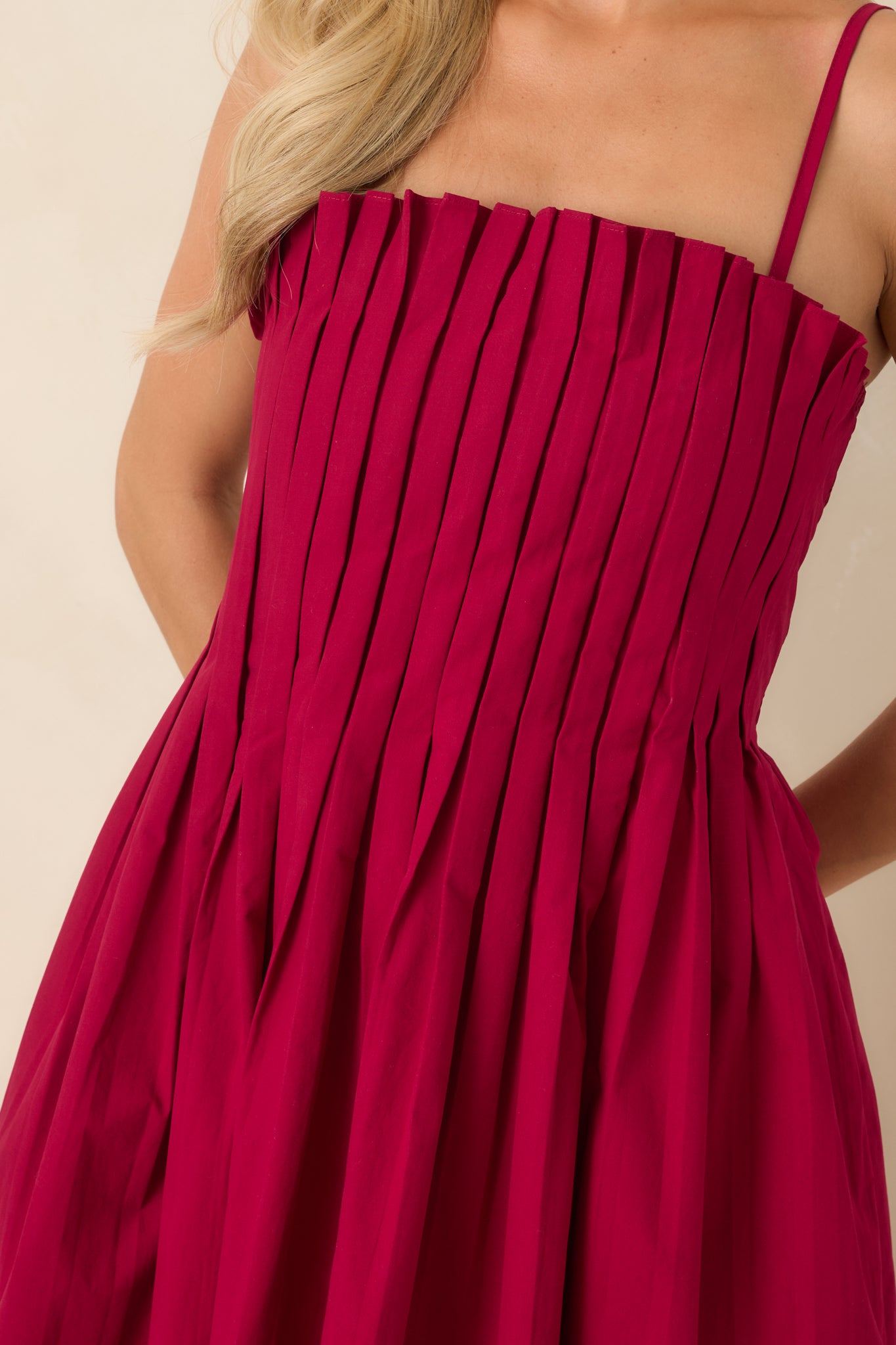 Close-up of the ruby red dress highlighting the square neckline, adjustable spaghetti straps, crisp vertical pleating across the fitted bodice, and the smooth cotton fabric.