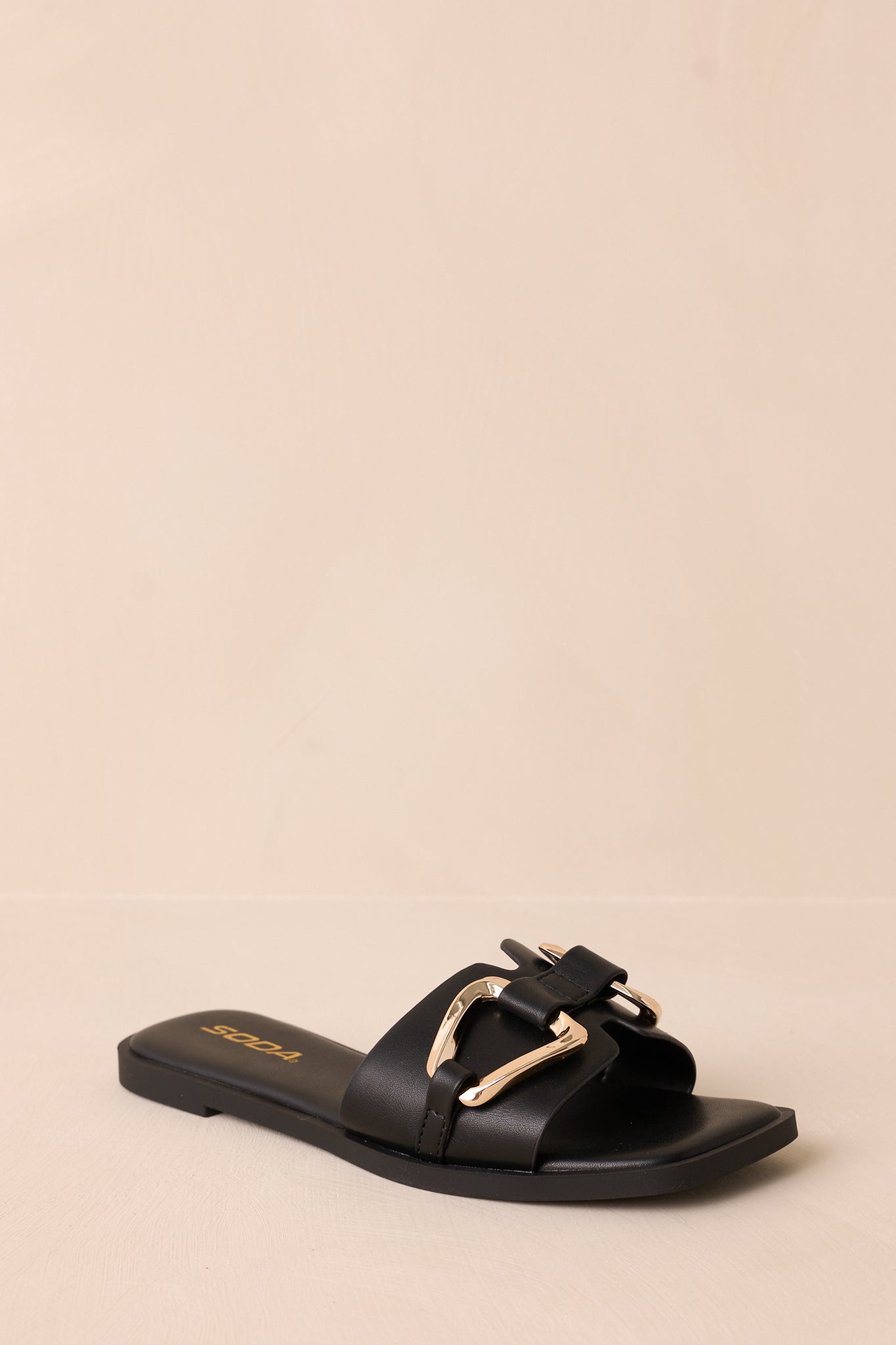 A slightly angled photo of the sandal showcasing its square toe, notched band, and the gold buckle, with a soft beige background.