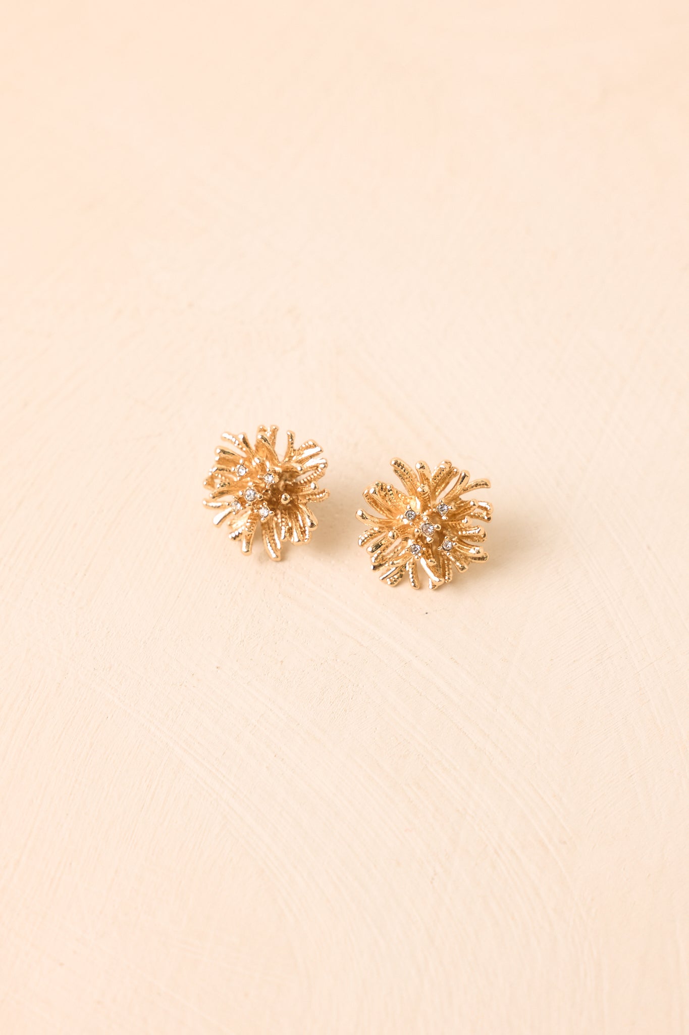 Sculptural gold-tone earrings against a beige background showcasing organic curves, textured strands, and sparkling crystal accents.
