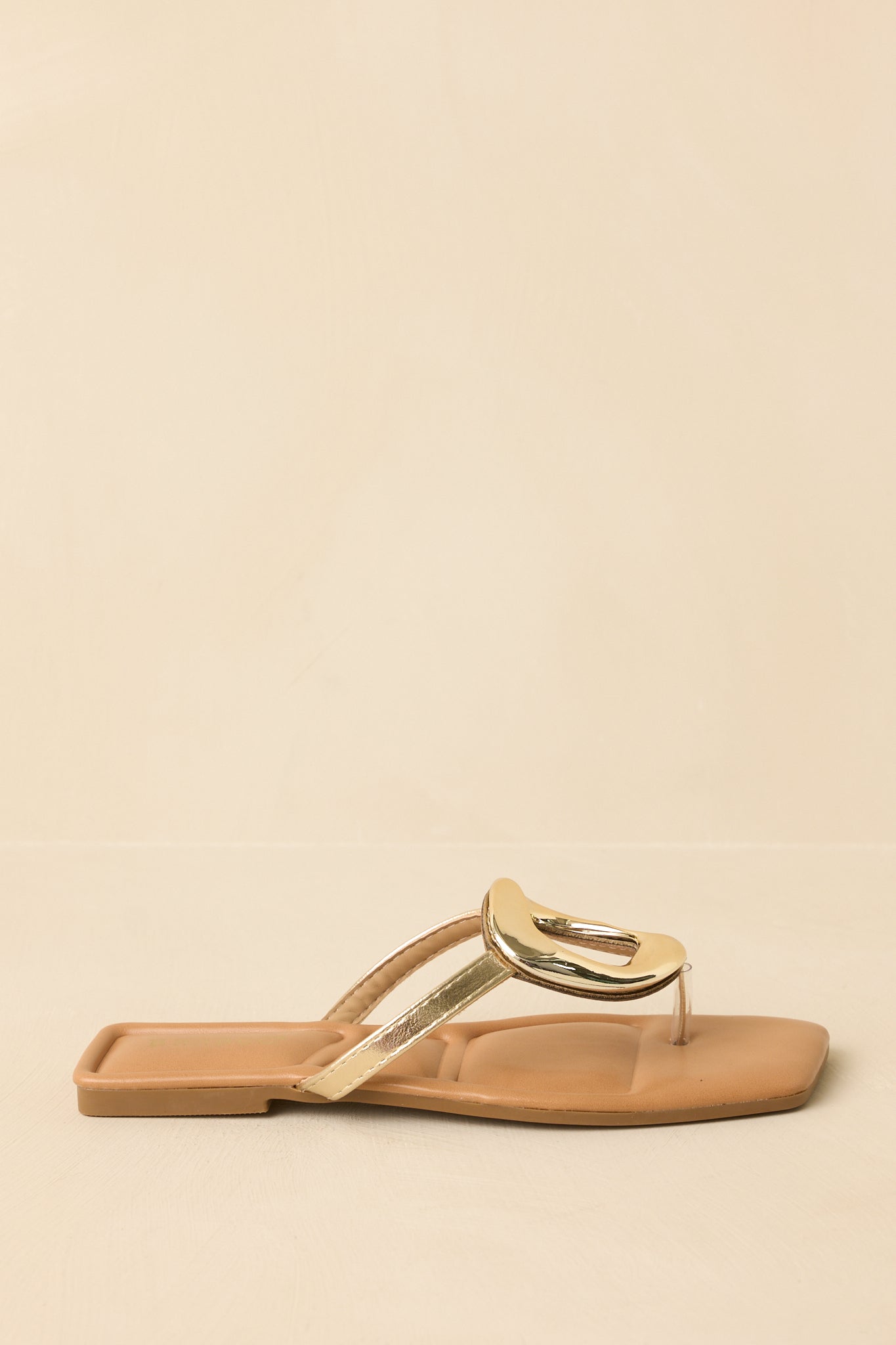 Side profile of the gold metallic sandals displayed against a beige background emphasizing the flat sole.