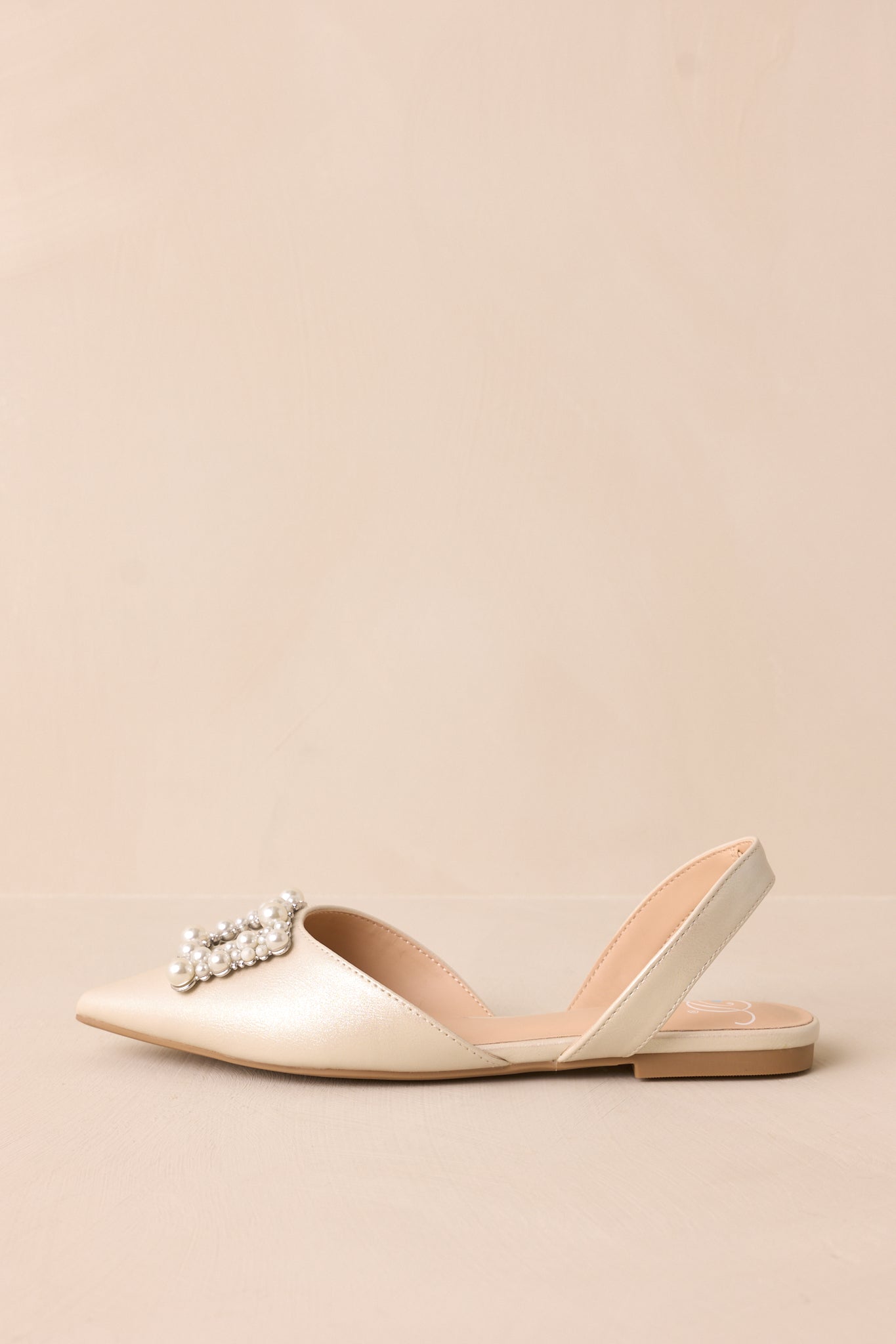 Full side view of the flats against a beige background, focusing on the pointed toe and large rhinestone square-shaped buckle with faux pearls.