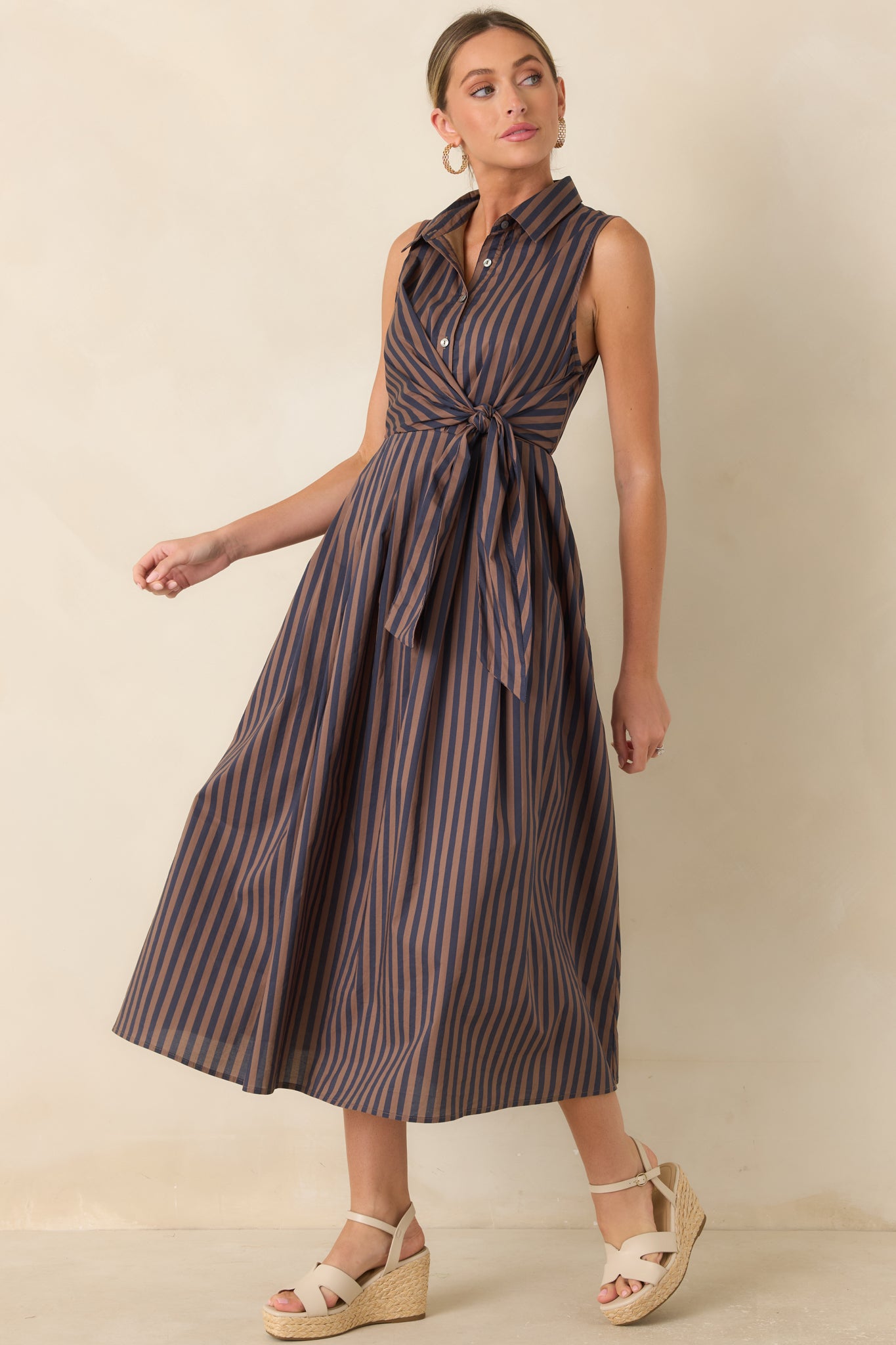 Sleeveless midi dress with vertical brown and navy stripes, a wrap-style tie front, side pockets, and a graceful silhouette.