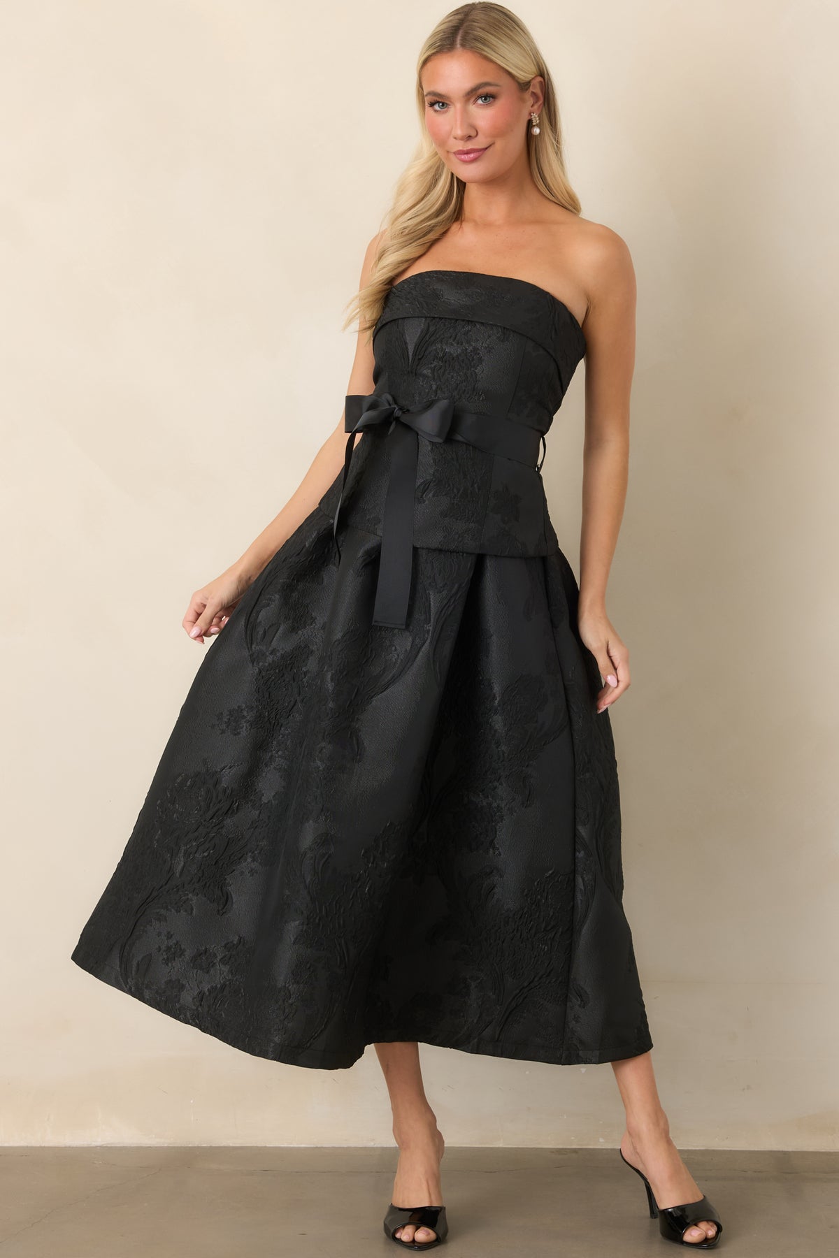 Structured strapless dress featuring a dramatic waist bow, split peplum overlay, and flowing pleated midi skirt.