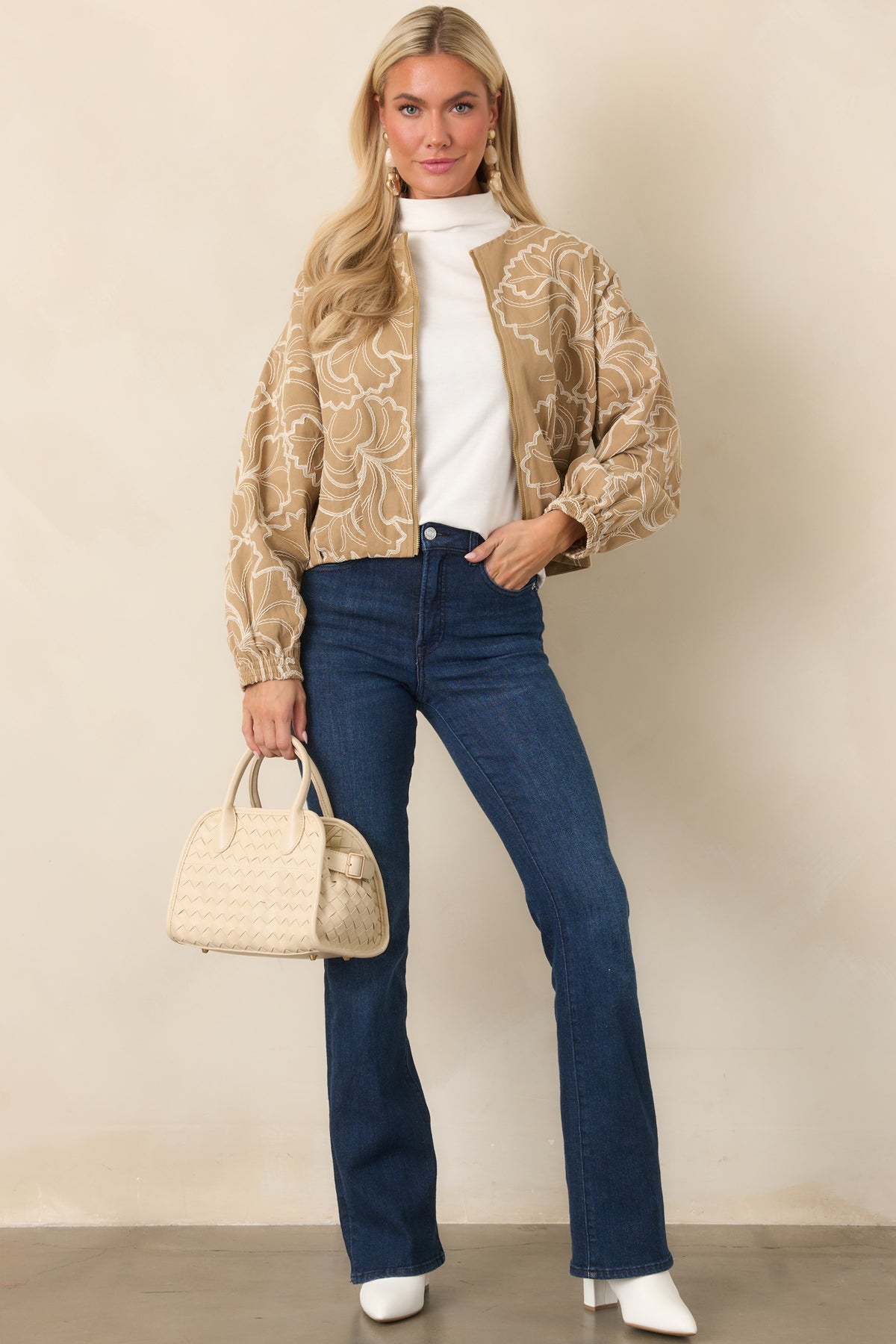 Relaxed tan jacket showcasing delicate ivory floral embroidery, polished gold-tone zipper hardware, and gathered sleeve cuffs.