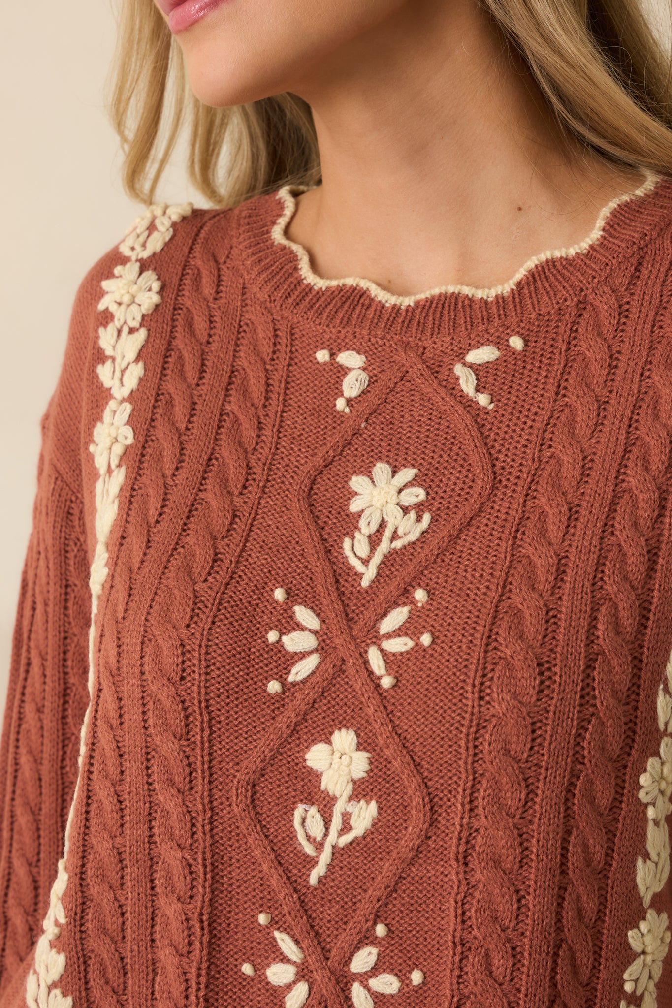 Up-close view of the terracotta cable knit showcasing the ivory embroidered flowers and scalloped trim