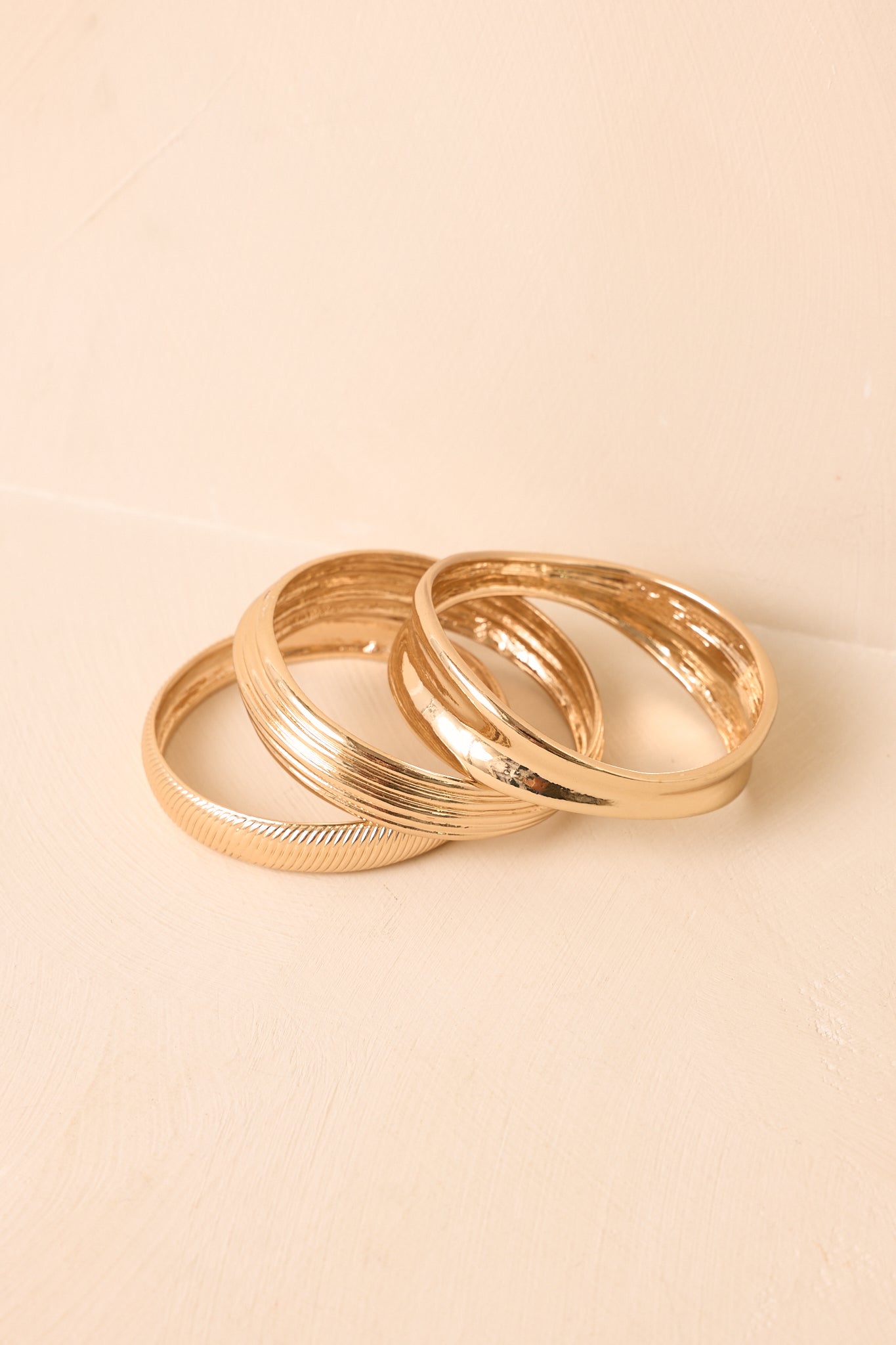 Three-piece gold-tone bangle set displayed against a beige background, highlighting the mixed finishes and dimensional design.
