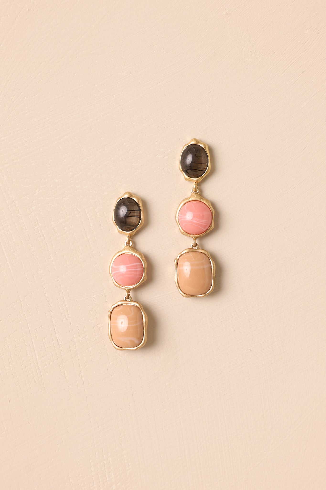 Three-stone drop earrings displayed on a beige background, showcasing the smoky brown, blush pink, and beige cabochons in gold settings.