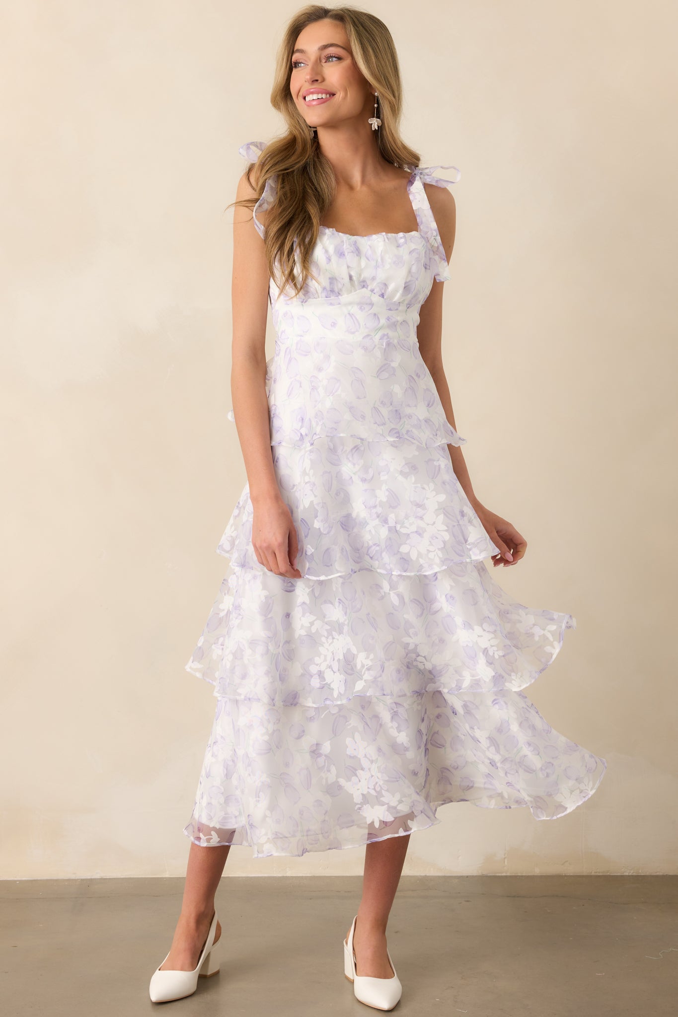 A tiered midi dress with a lavender tulip floral tulle overlay, self-tie bow straps, and a soft white lining. The dress moves gracefully, highlighting its airy fabric.