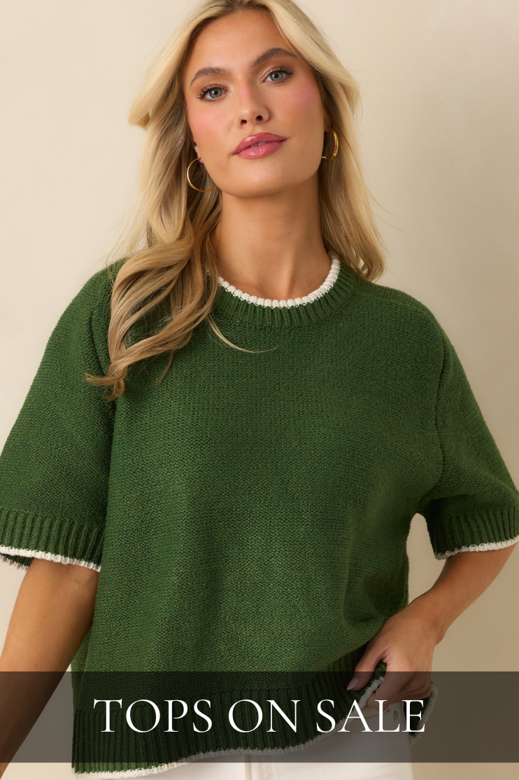 Woman wearing a green knit short-sleeve sweater with white trim and gold hoops, showcasing cozy and casual pieces from the Tops on Sale collection.