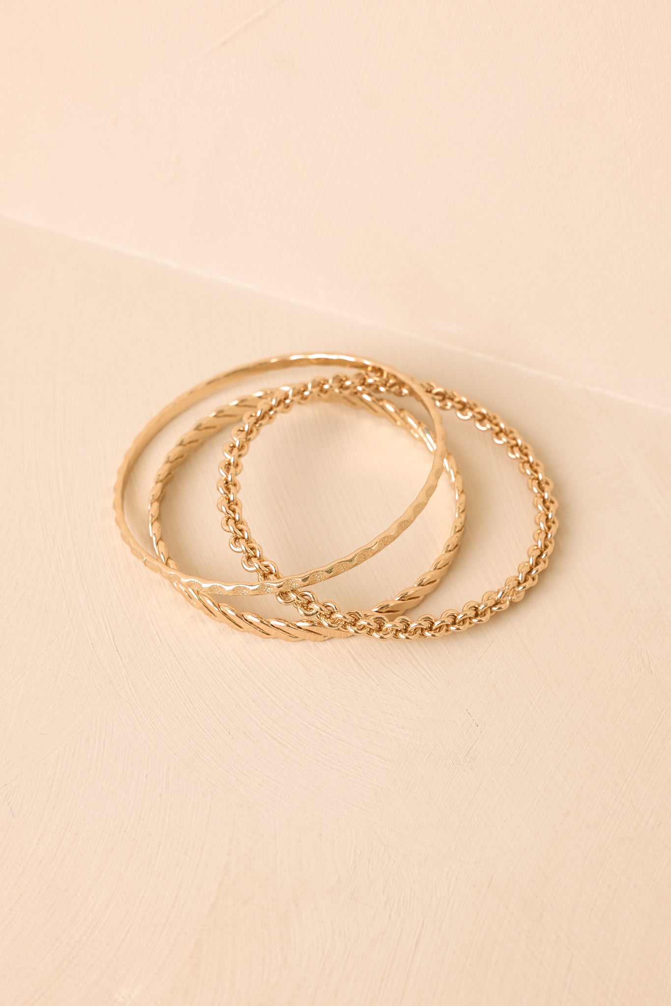 Trio of lightweight gold bangles showcasing mixed textured details displayed against a beige background.