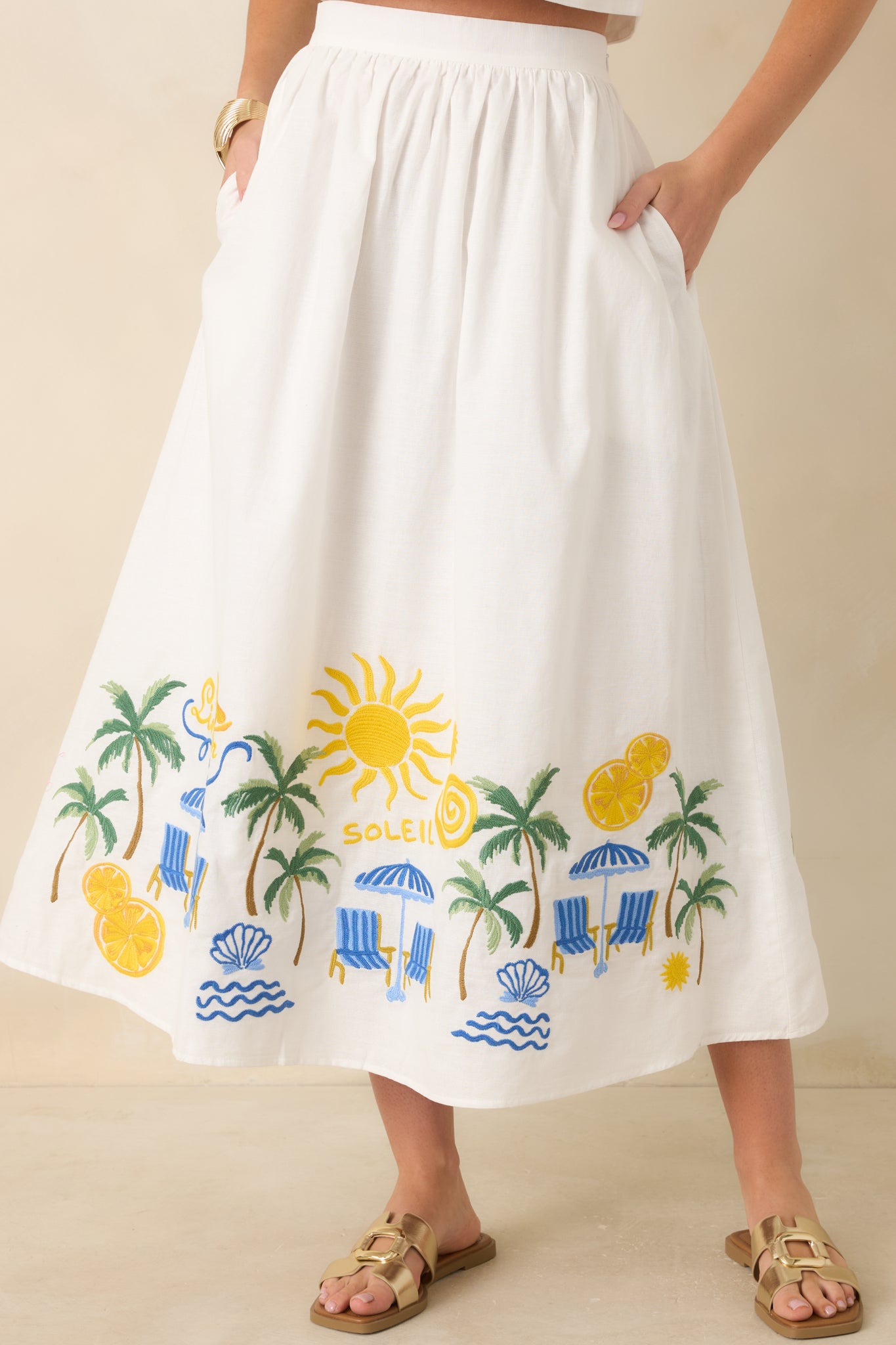 Detail view of the embroidered hem, showcasing sun, palm, and seaside motifs with vibrant stitching against the crisp white fabric.