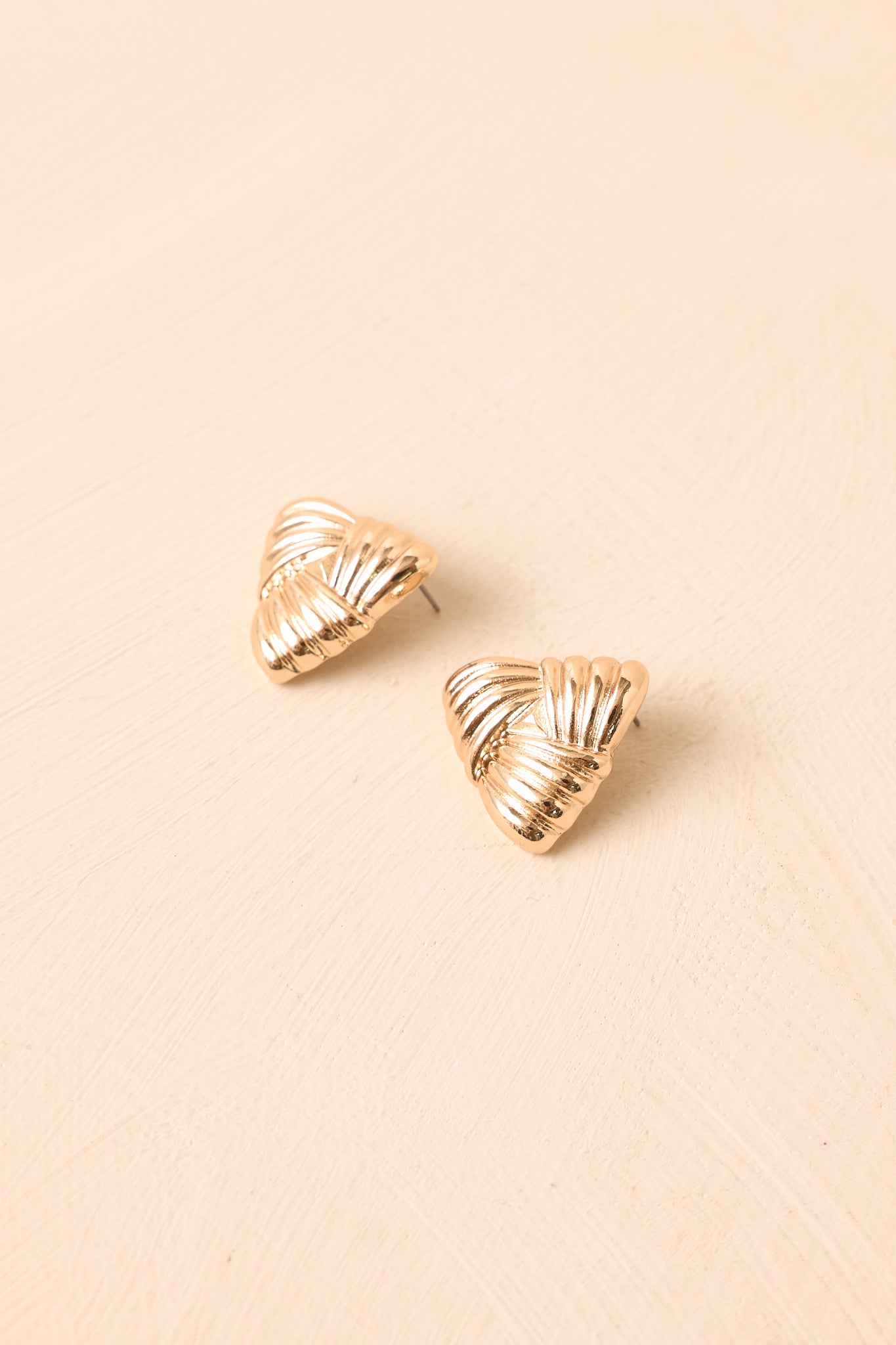 Gold-tone sculptural stud earrings with layered, woven-inspired folds presented on a neutral beige backdrop