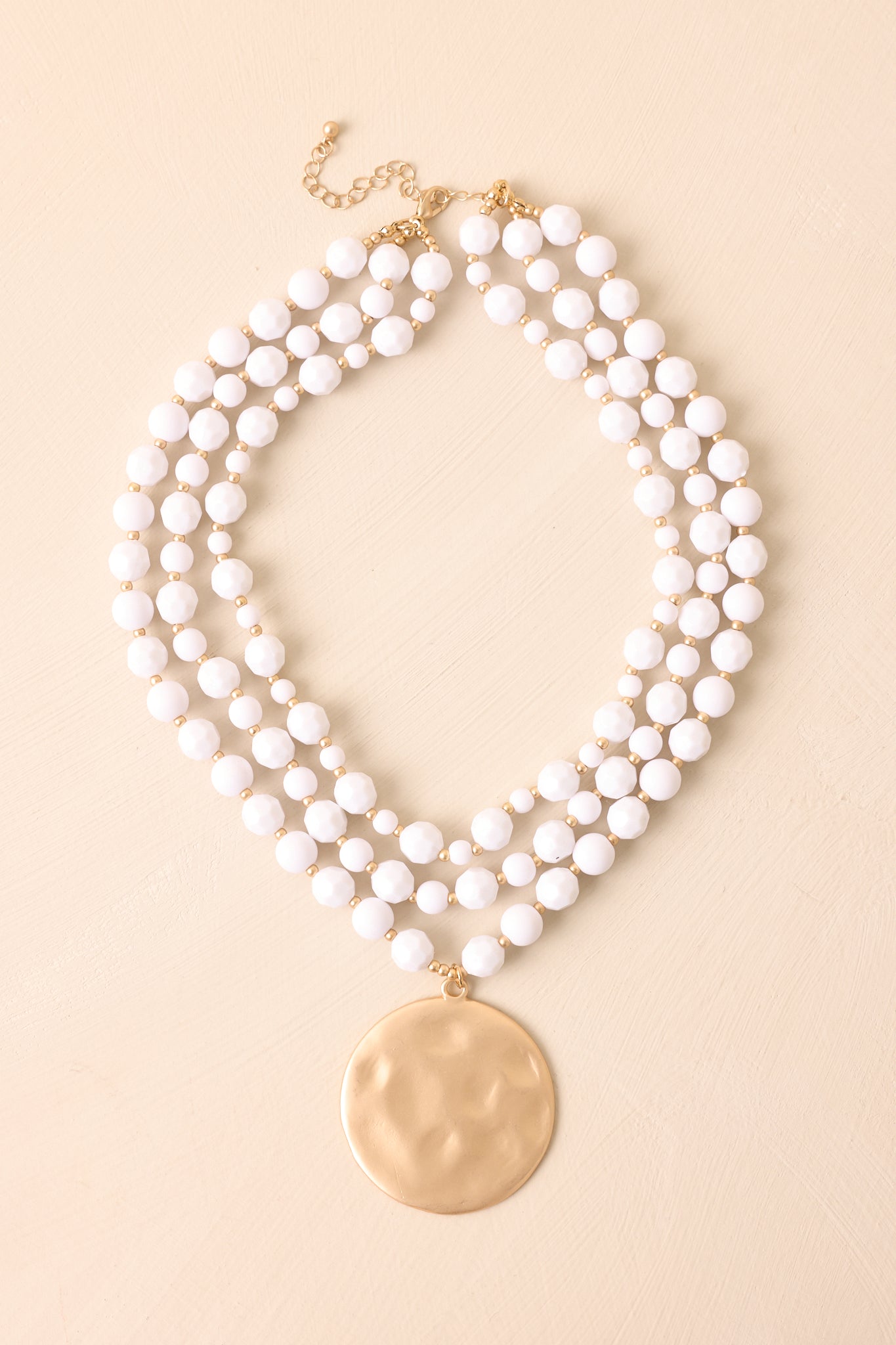 The white and gold beaded layered necklace displayed on a beige background with a hammered disc pendant.
