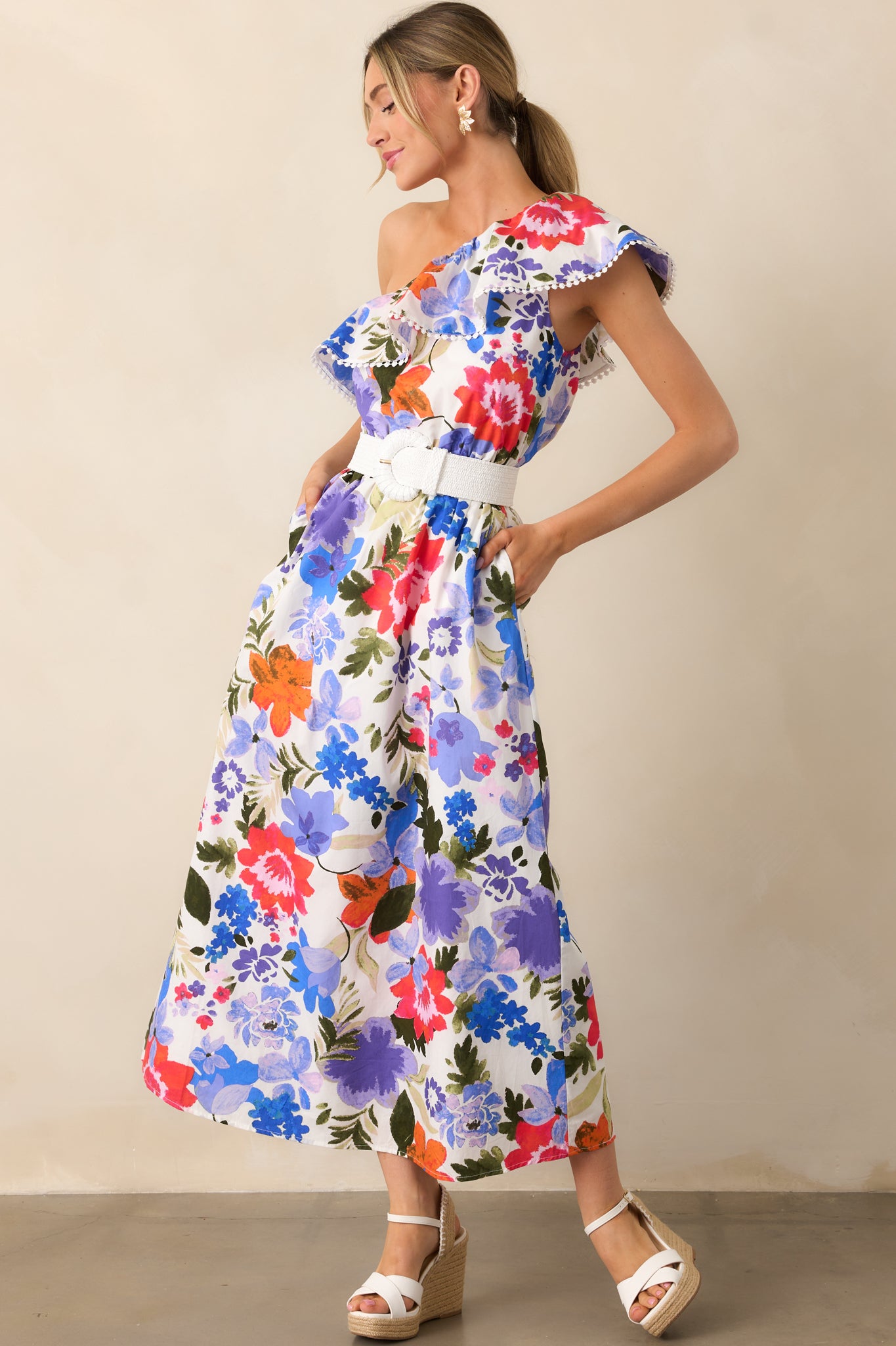 A full view of the maxi dress with a white background featuring a blue and red floral print, an elastic one-shoulder ruffle neckline, and functional pockets for a stylish, feminine look.