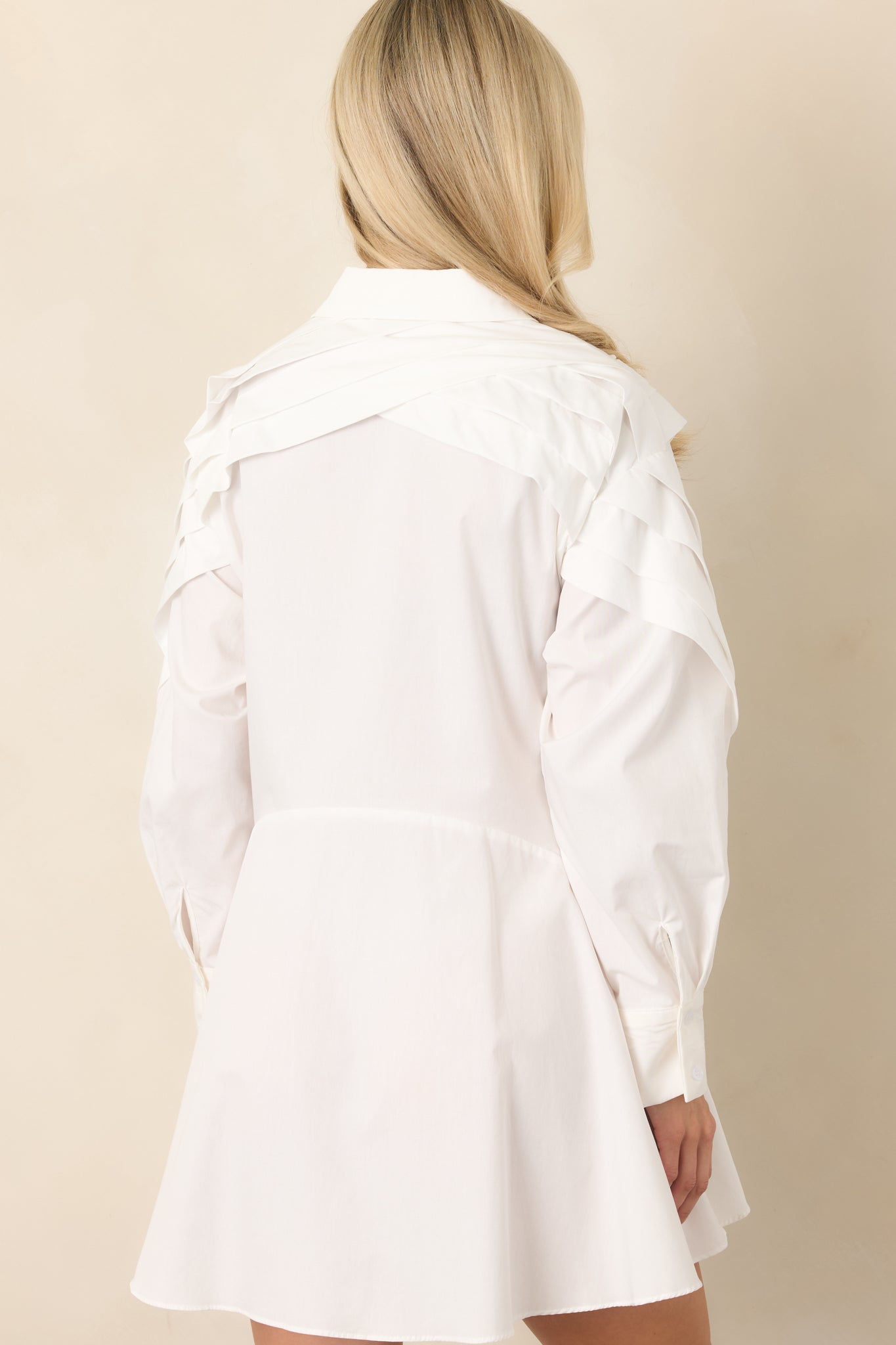 Rear view of the white woven cotton mini dress, highlighting the layered pleated yoke, long sleeves with button cuffs, and gently flared skirt.
