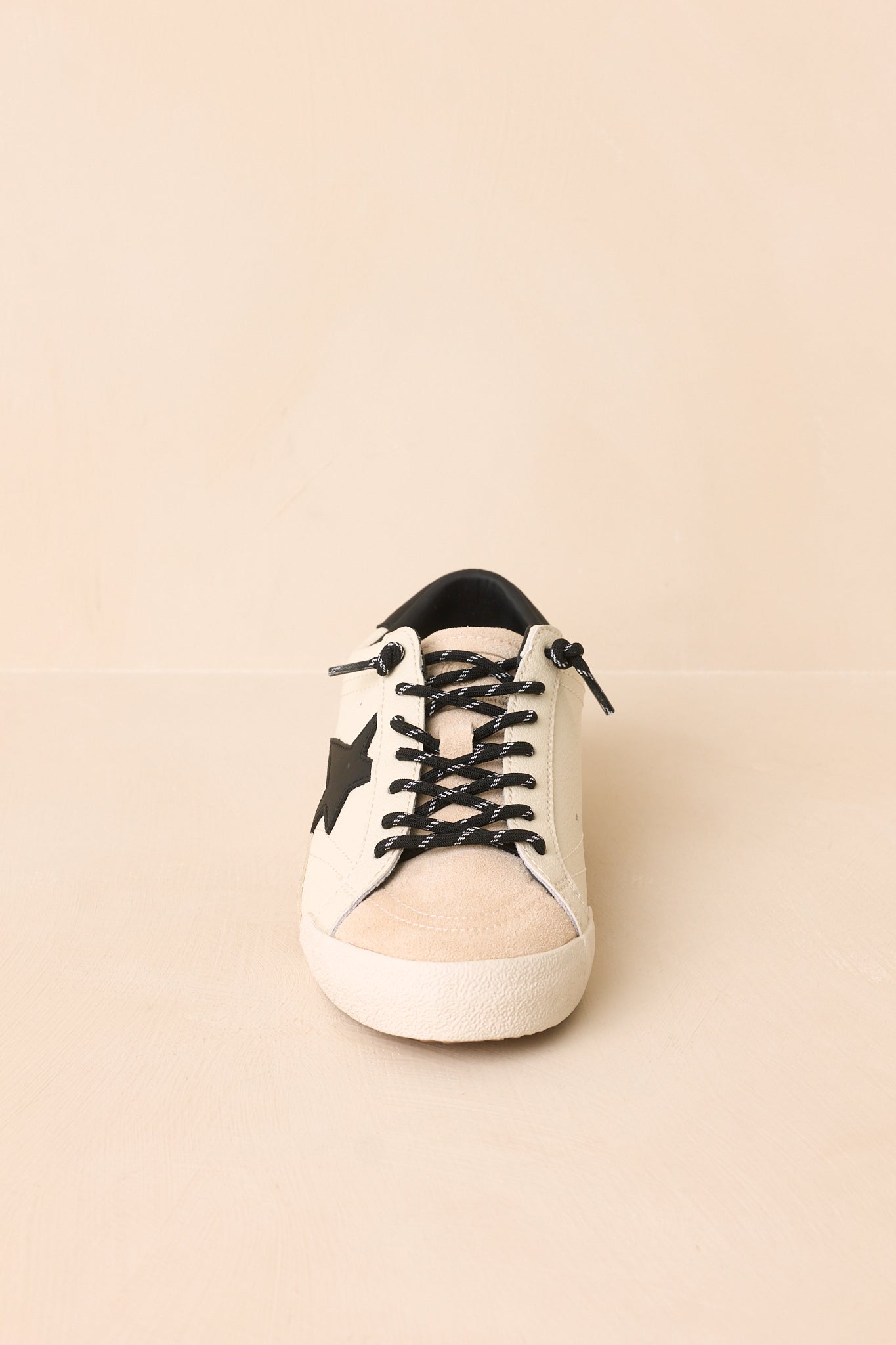White statement sneakers with a signature black star, black heel detail, layered textures, and a classic low-profile design displayed on a beige background.