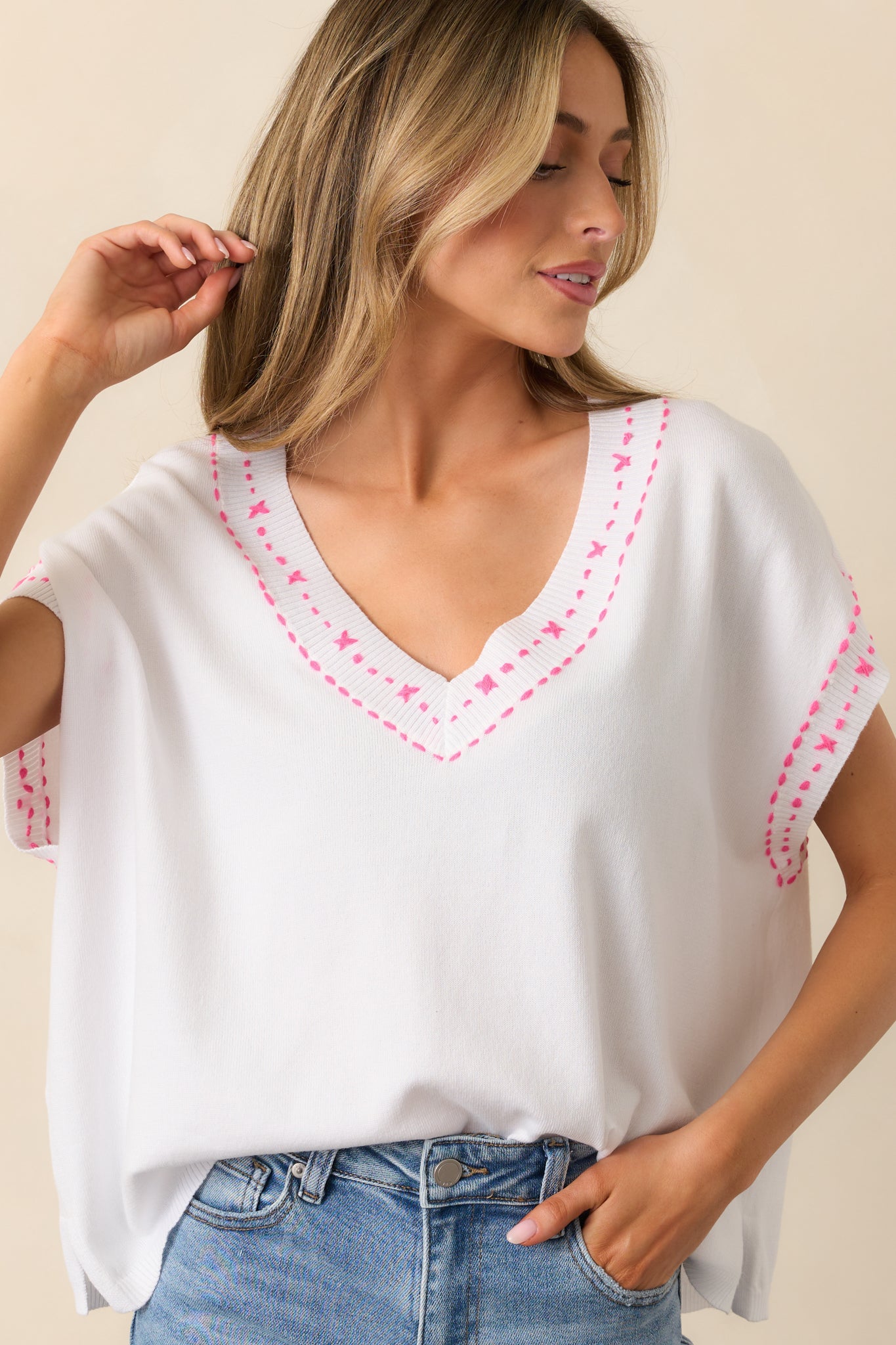 White top featuring a V-neckline, pink embroidered stitching on the neckline and sleeves, a relaxed fit, and batwing sleeves.