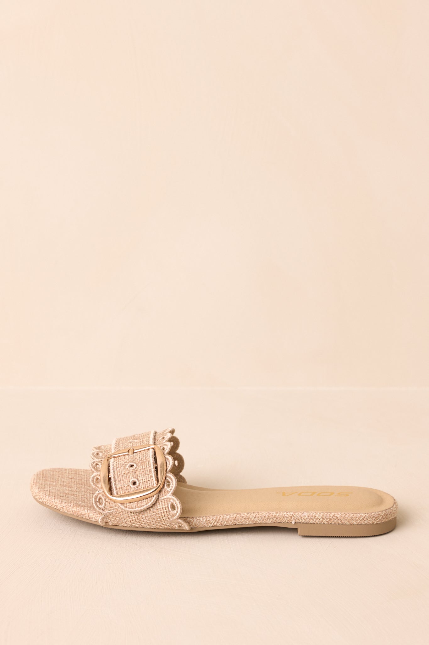Side view of woven slip-on sandals highlighting scalloped edges and buckle hardware on beige surface.