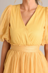 Close up view of this yellow dress features a v-neckline, puff sleeves, elastic stretch around the waist, a removable self-tie at the waist, and a tiered flowy skirt.