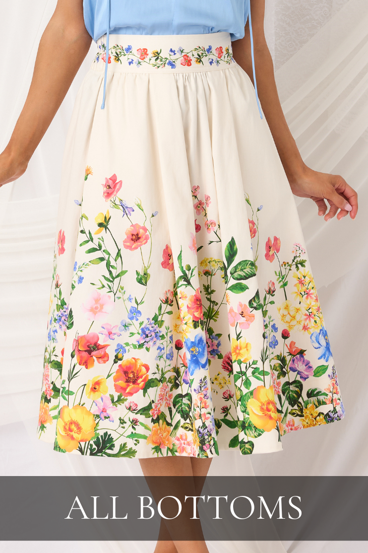 Model wearing a cream midi skirt with colorful floral garden print and a light blue top, highlighting the women’s bottoms collection with bright spring patterns and feminine styling.