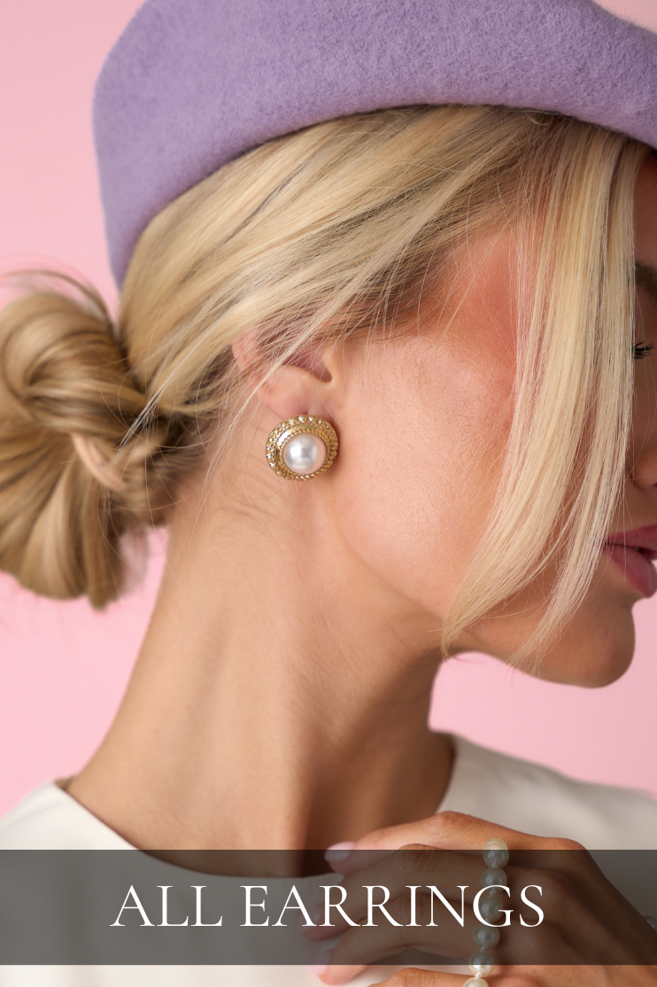 Close-up of gold and pearl stud earrings styled with a beret, linking to the All Earrings shop.