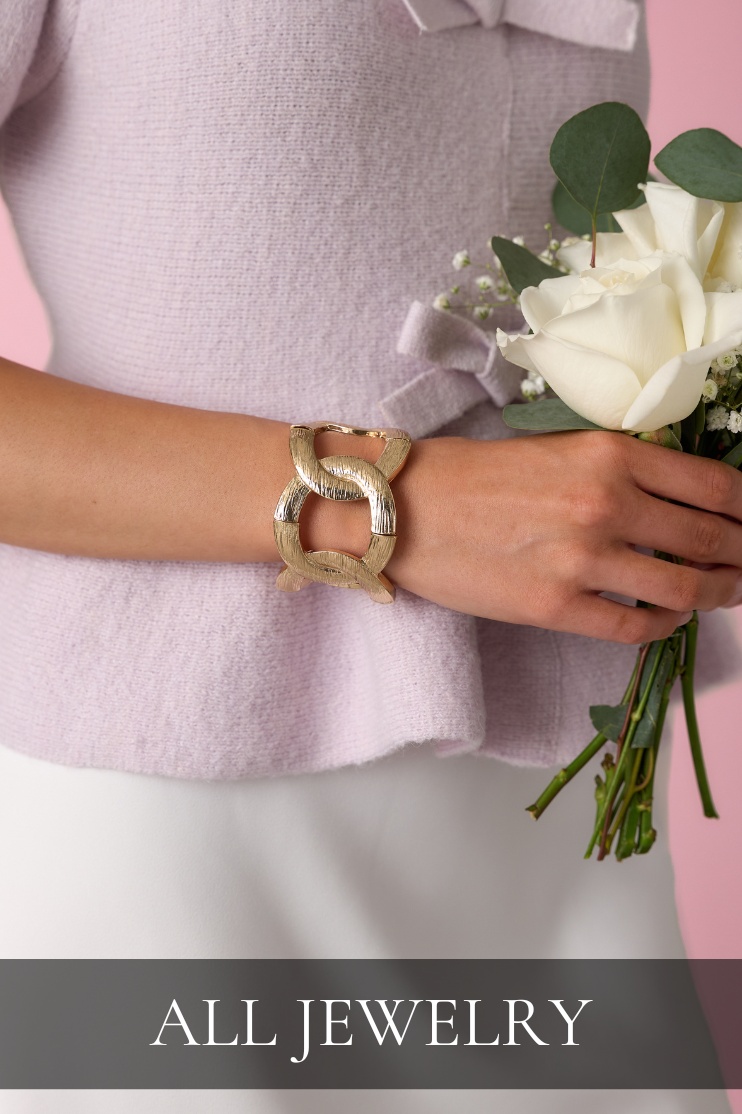 Close-up of a chunky gold bracelet styled with a soft knit top and white flowers, linking to the All Jewelry shop.
