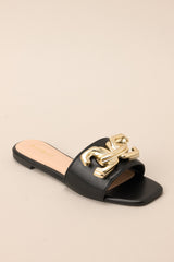 Front angled view of these black flat sandals that feature a slip on style, and a large gold buckle-like design.
