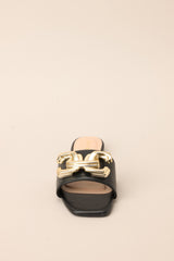 Front view of these black flat sandals that feature a slip on style, and a large gold buckle-like design.