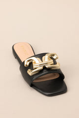 Close up front view of these black flat sandals that feature a slip on style, and a large gold buckle-like design.