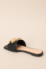 Back angled view of these black flat sandals that feature a slip on style, and a large gold buckle-like design.