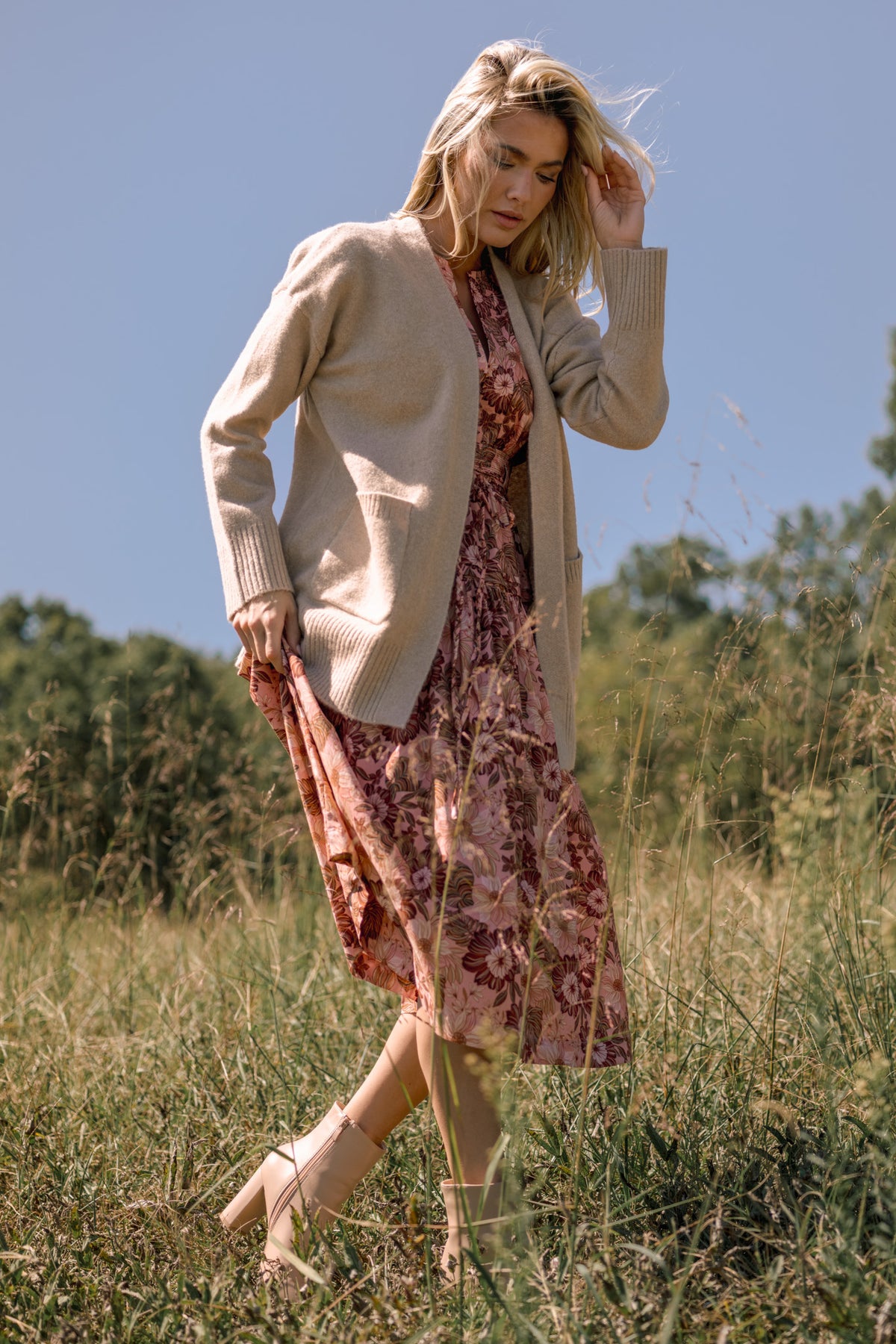 Woman wearing a beige open-front cardigan over a pink floral midi dress with neutral ankle boots in a grassy field.