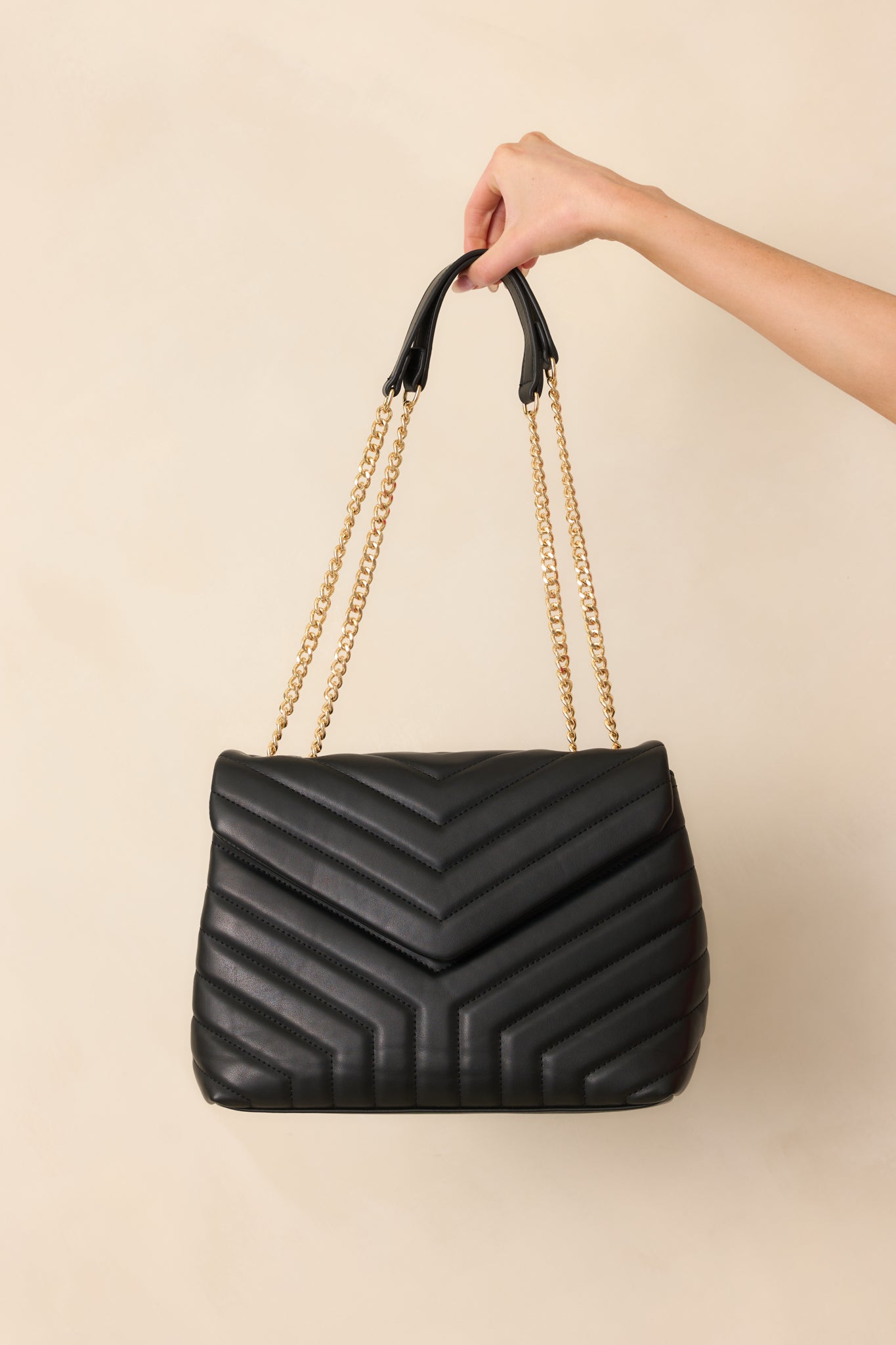 A black quilted rectangular handbag with a snap flap closure and black zipper closure, featuring two gold chain handles with black faux leather handles.