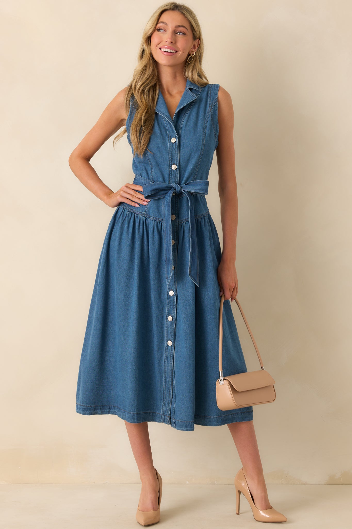 The blue denim sleeveless dress shown front facing with a drop waist and adjustable tie belt.