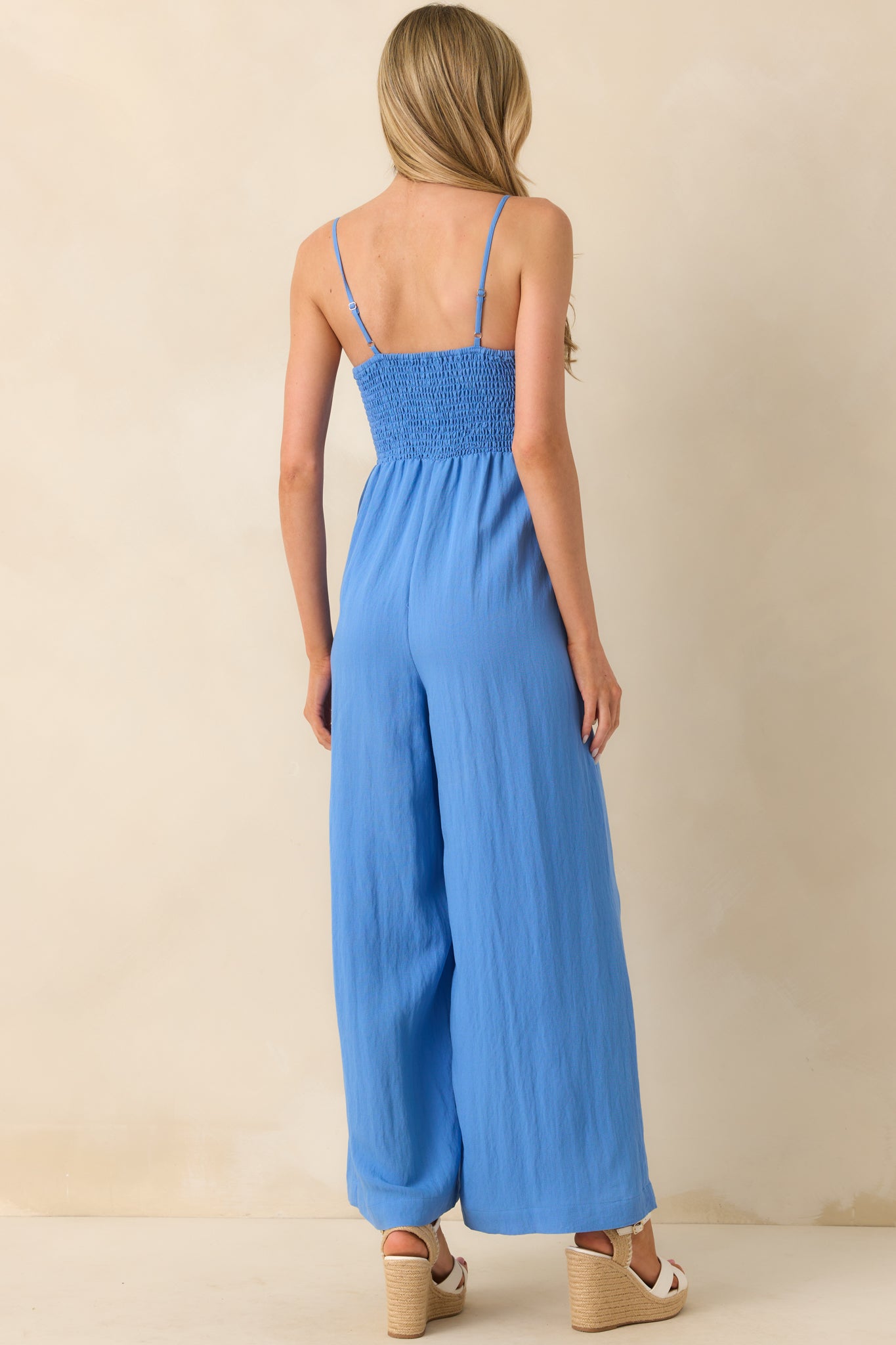 Photo of back of model wearing a ivory jumpsuit that features a fully smocked bust, adjustable spaghetti straps, lightweight rayon blend fabric, and wide ankle length legs.