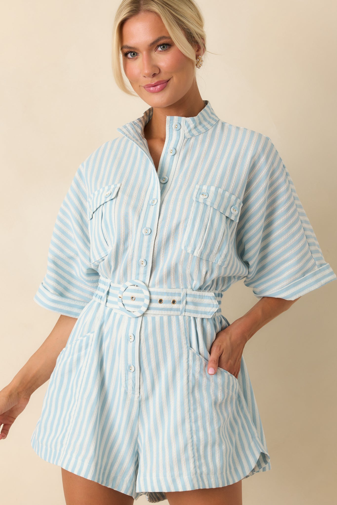 Close-up of light blue striped romper featuring detailed stitching, collared neckline, button front, cuffed short sleeves, belted waist, and front patch pockets.